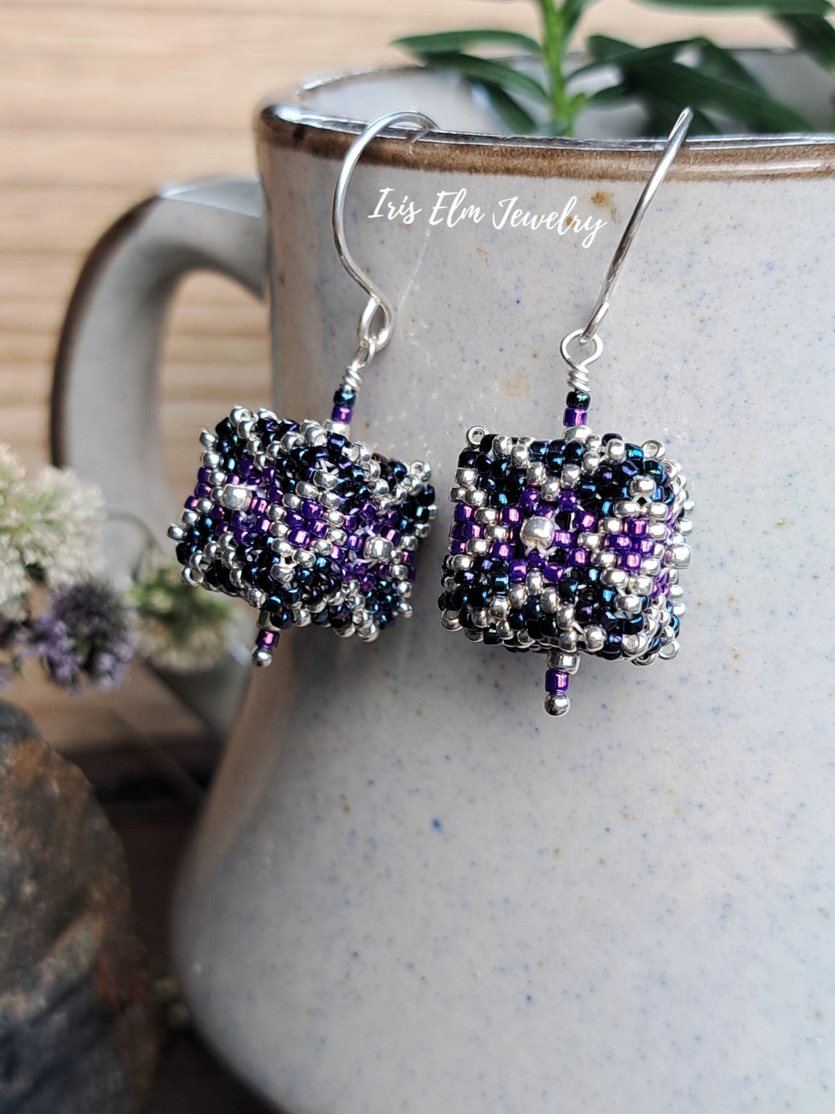 A pair of handmade beaded dangle earrings by Iris Elm Jewelry, featuring purple and black seed beads wrapped in silver wire to form a textured cube shape, hanging from silver fishhook wires in front of a ceramic mug.
