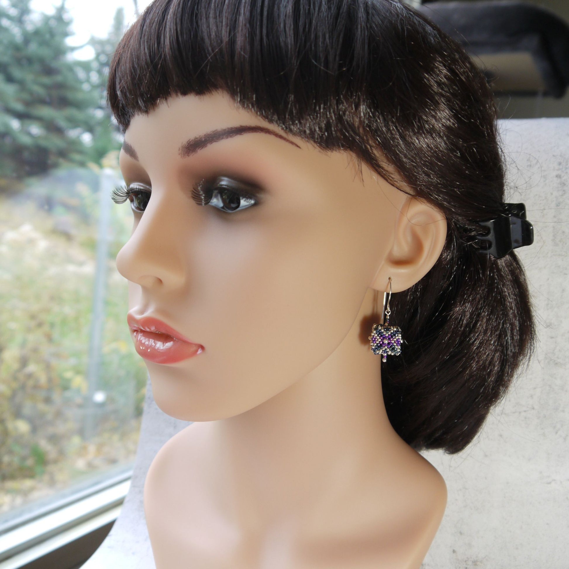 Mannequin head with dark hair styled in a ponytail, wearing a purple and blue cube earring.