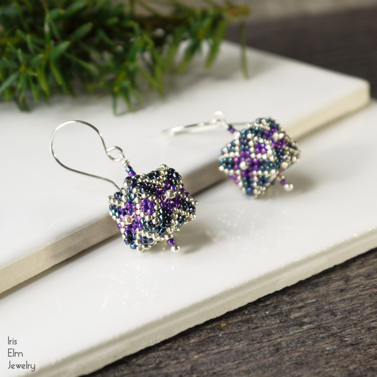 Beadwoven silver, blue, and purple cube shaped seed bead glass earrings on a white surface with a plant in the background