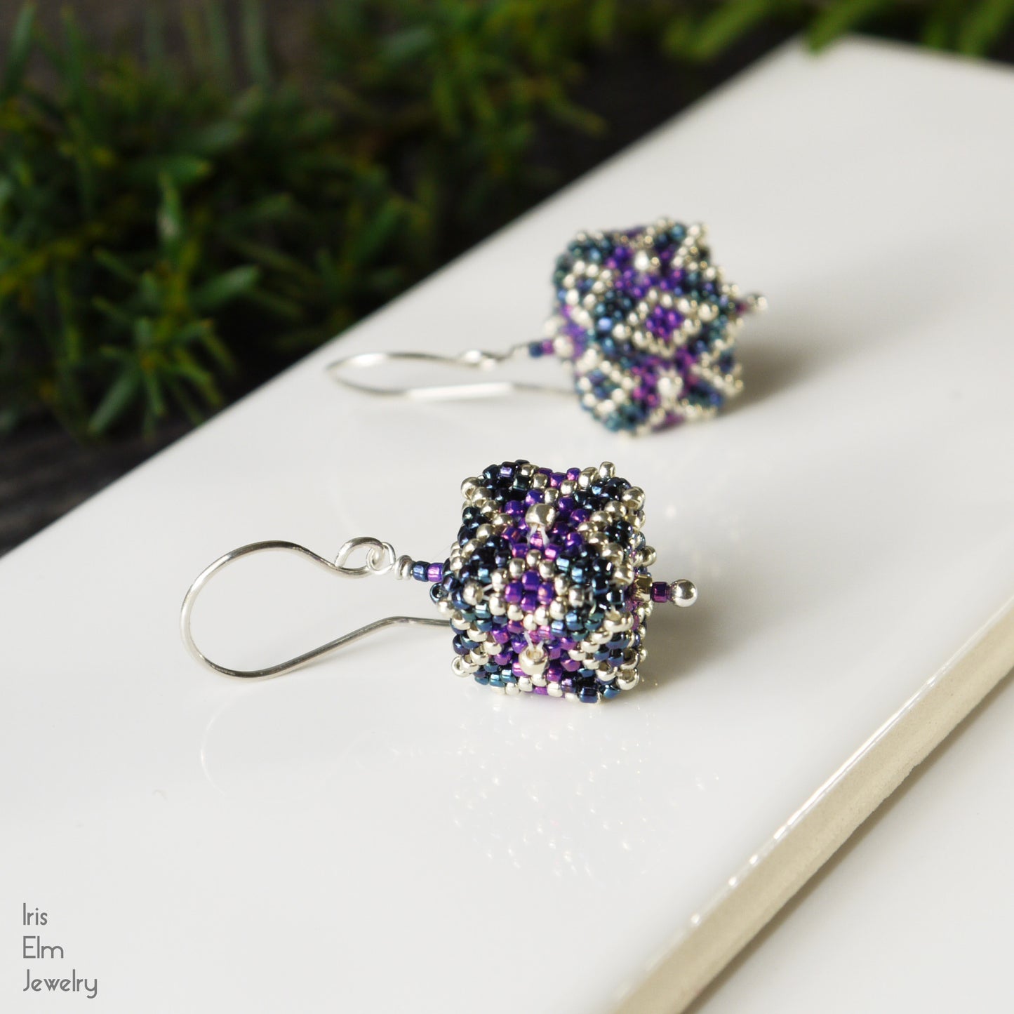 Pair of beaded cube seed bead earrings on a white surface with a blurred green background