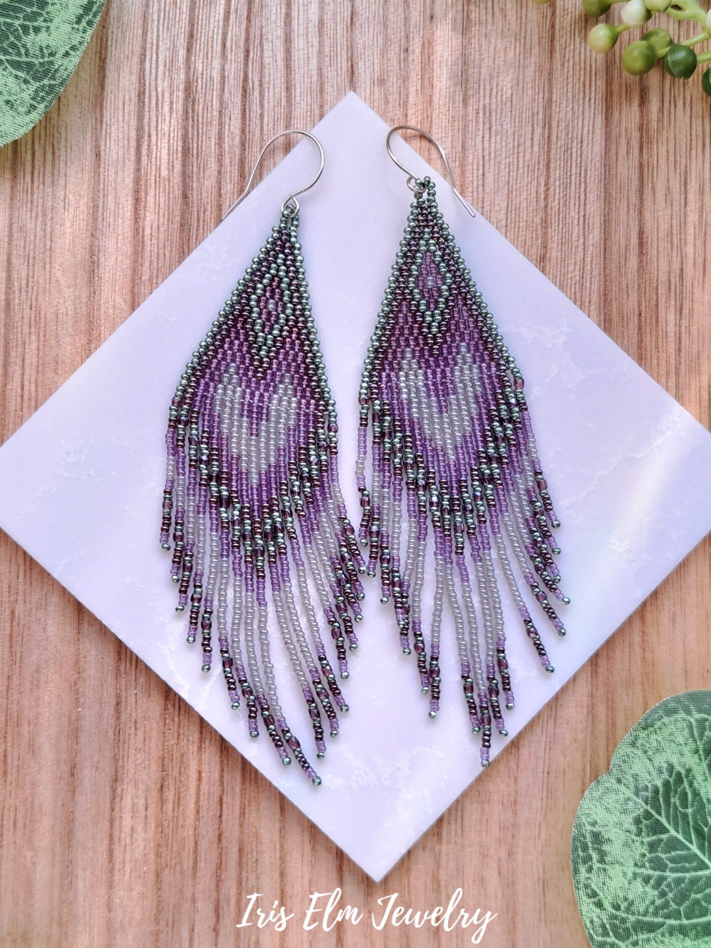 Purple green grey seed bead beaded fringe earrings on a white square with 'Iris Elm Jewelry' branding, placed on a wooden surface.