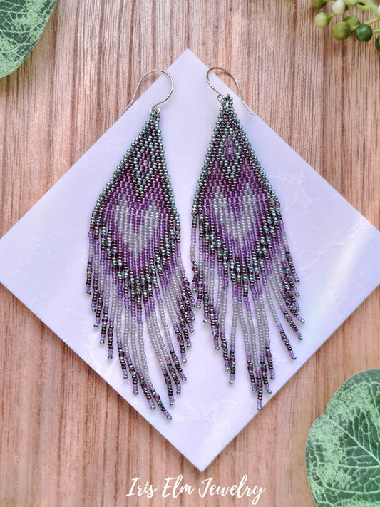 Purple green grey seed bead beaded fringe earrings on a white square with 'Iris Elm Jewelry' branding, placed on a wooden surface.
