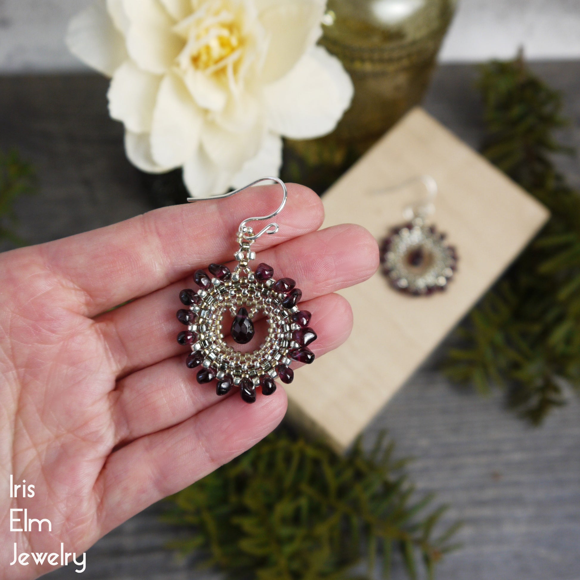 Hand holding a beaded red garnet gemstone and golden glass seed bead earring with a blurred background featuring another earring and decorative elements.