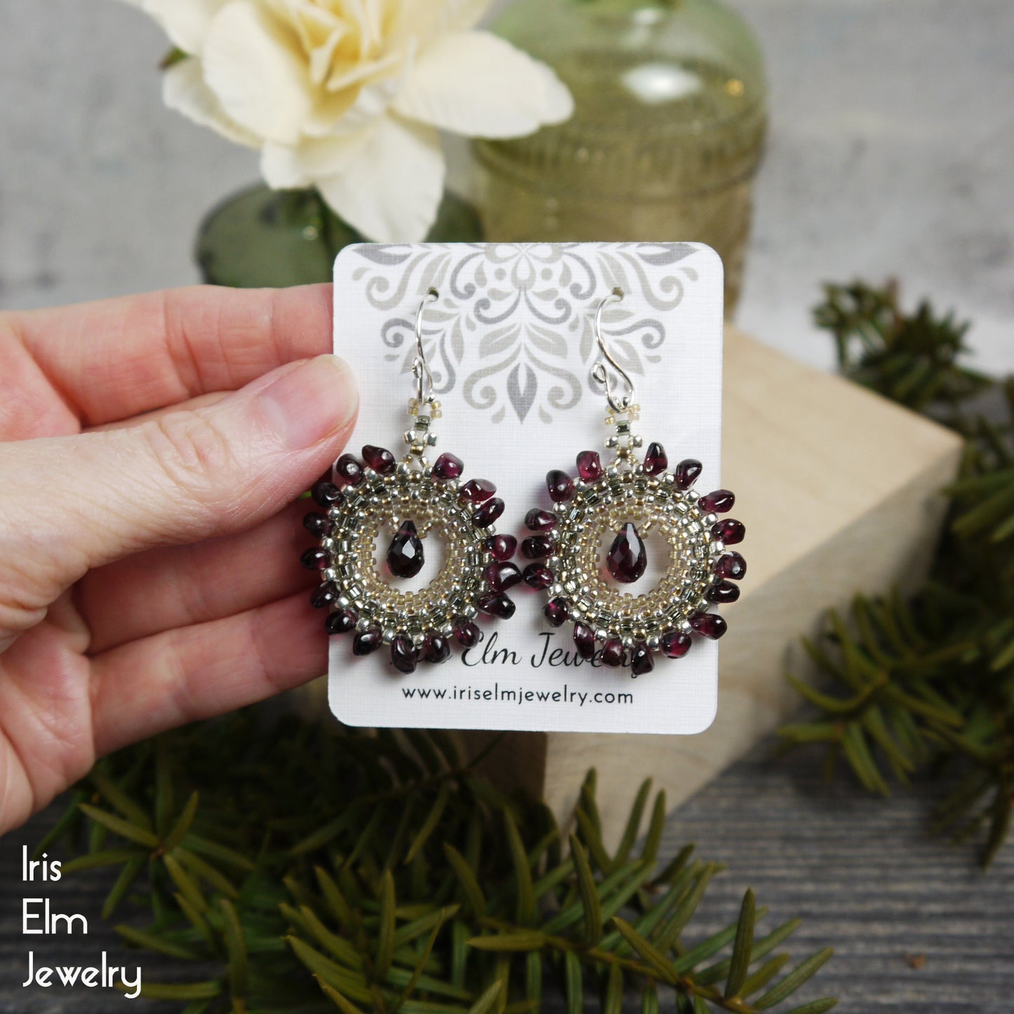 garnet beadwork Earrings on a card held by a hand with floral and greenery in the background