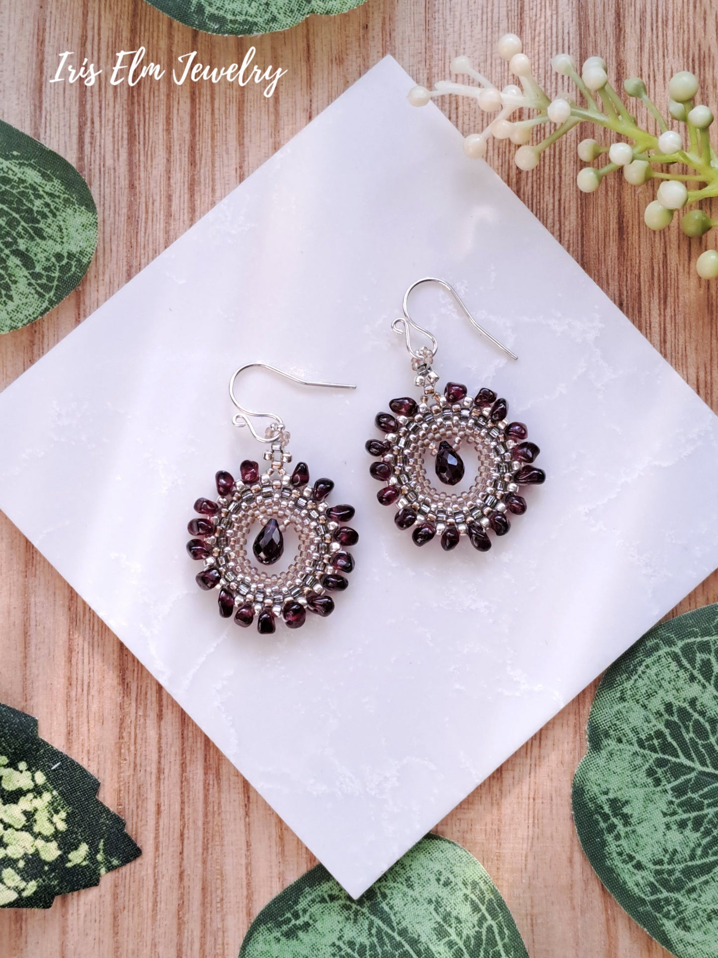 Pair of beaded garnet earrings on a white tile with green leaf patterns