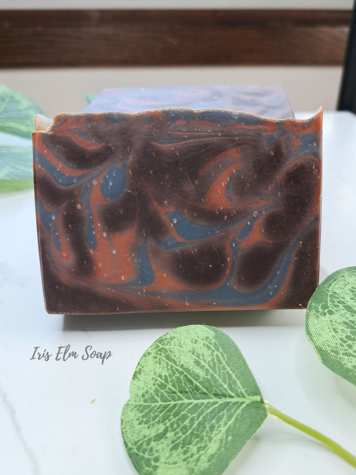A rectangular handmade cocoa butter cashmere soap bar with a marbled swirl pattern of dark brown, terracotta orange, and slate blue, displayed on a white marble surface with decorative green leaves. The brand name 'Iris Elm Soap' is printed in black script on the lower left.