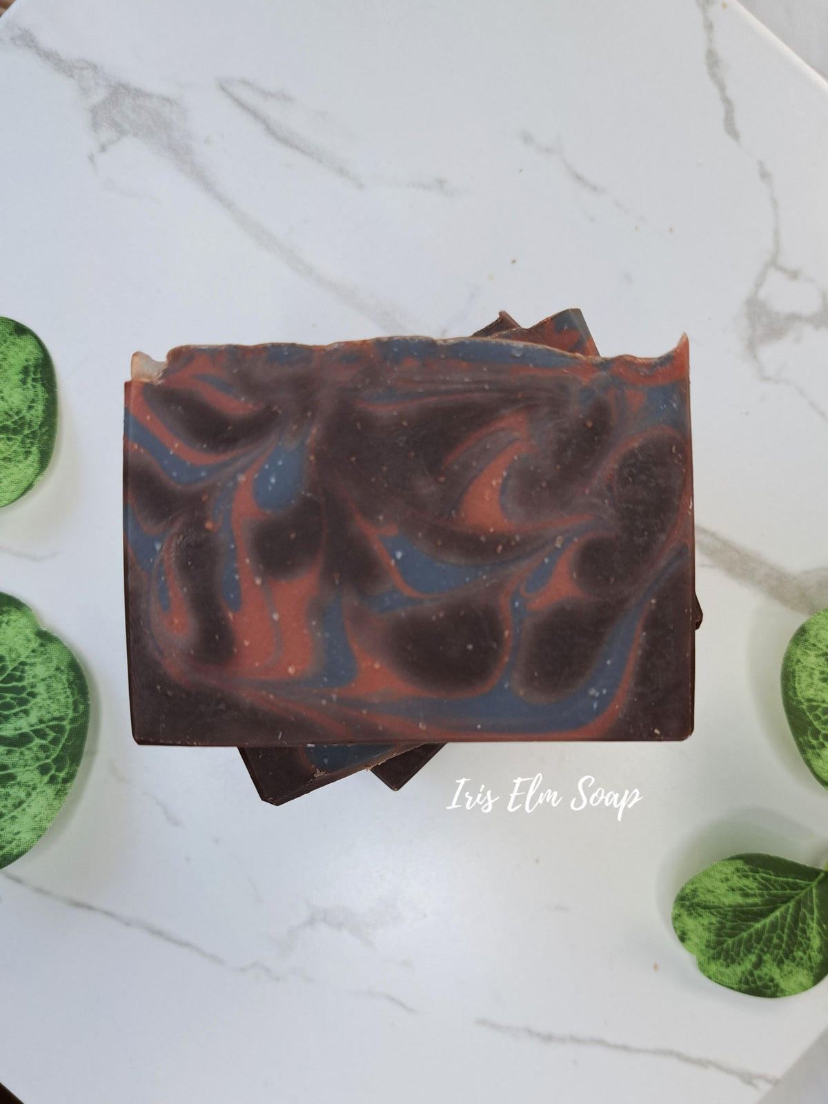 A rectangular bar of cocoa butter cashmere  artisan soap with a marbled swirl pattern of dark brown, rust red, and slate blue, displayed on a white marble surface with green decorative leaves.