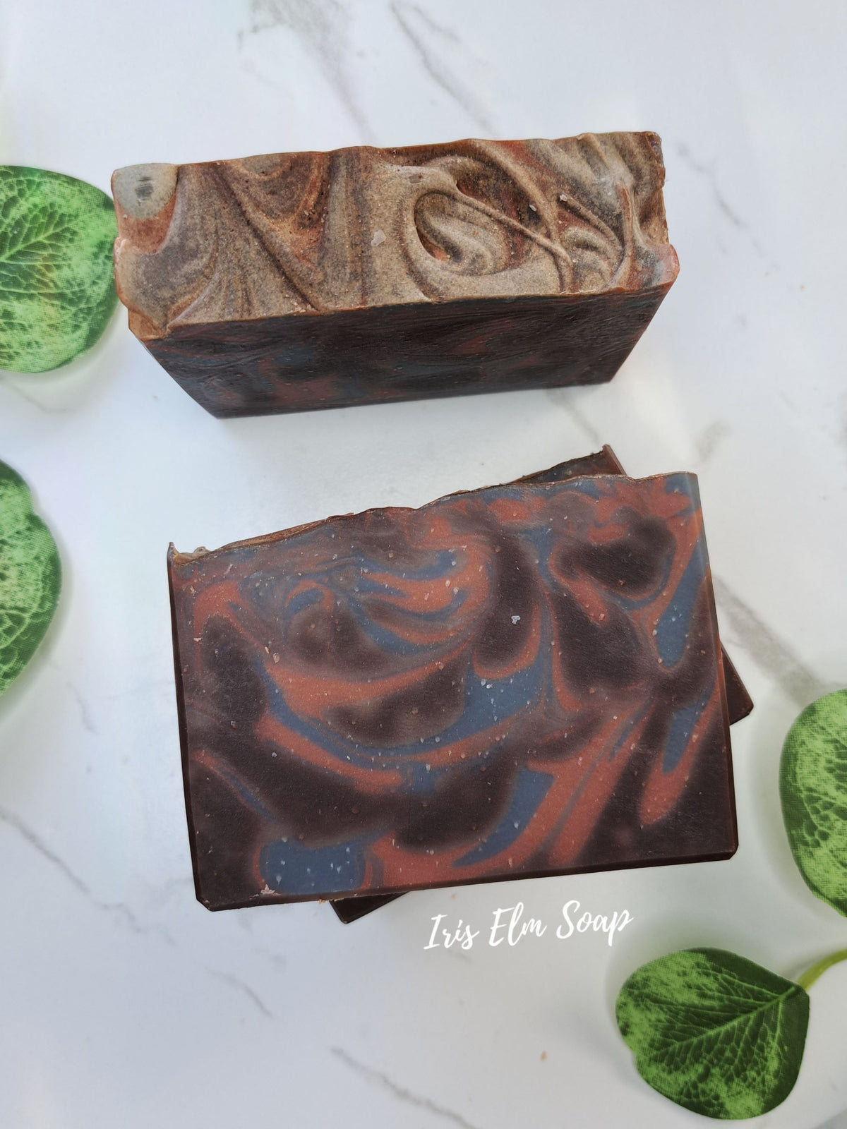 Two rectangular artisan cocoa butter cashmere soap bars with marbled swirls of dark brown, reddish-brown, and blue, styled on a white marble surface with green leaves. The brand name 'Iris Elm Soap' is printed on the front soap bar.
