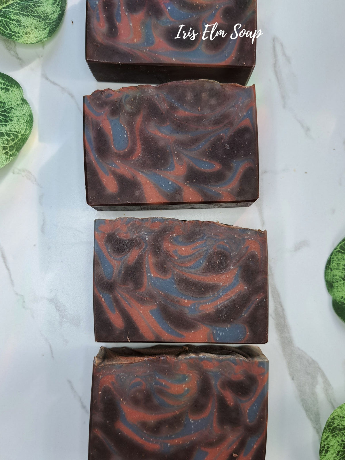 Four rectangular bars of cocoa butter cashmere artisanal soap with a marbled swirl pattern of dark brown, rust red, and slate blue, arranged vertically on a white marble surface with decorative green leaves at the corners. The text 'Iris Elm Soap' is visible in the top soap.
