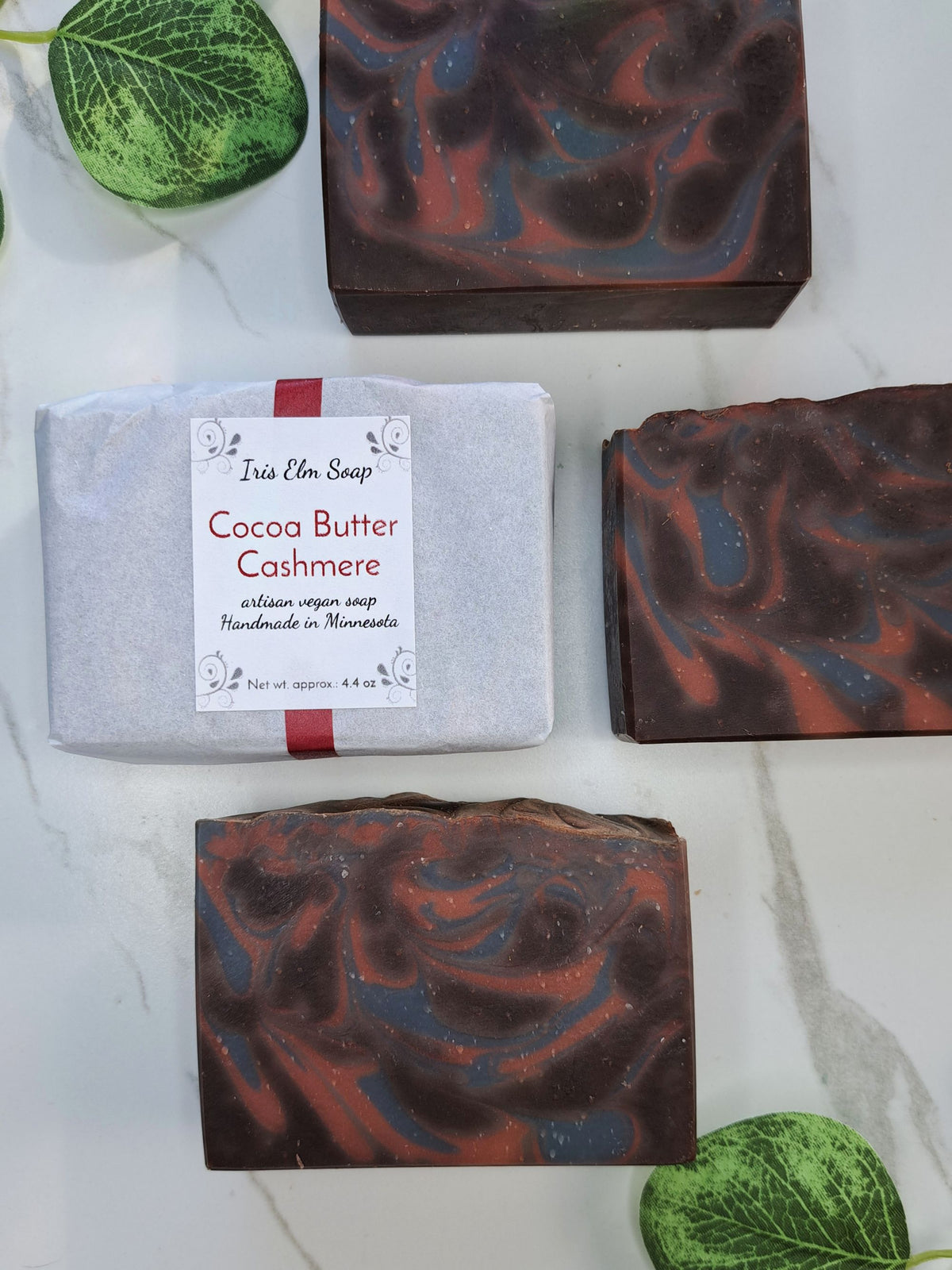 Four bars of artisan soap from Iris Elm Soap are displayed on a white marble surface, including one wrapped bar labeled 'Cocoa Butter Cashmere' and three unwrapped bars showing a marbled swirl pattern of dark brown, red, and blue-gray.