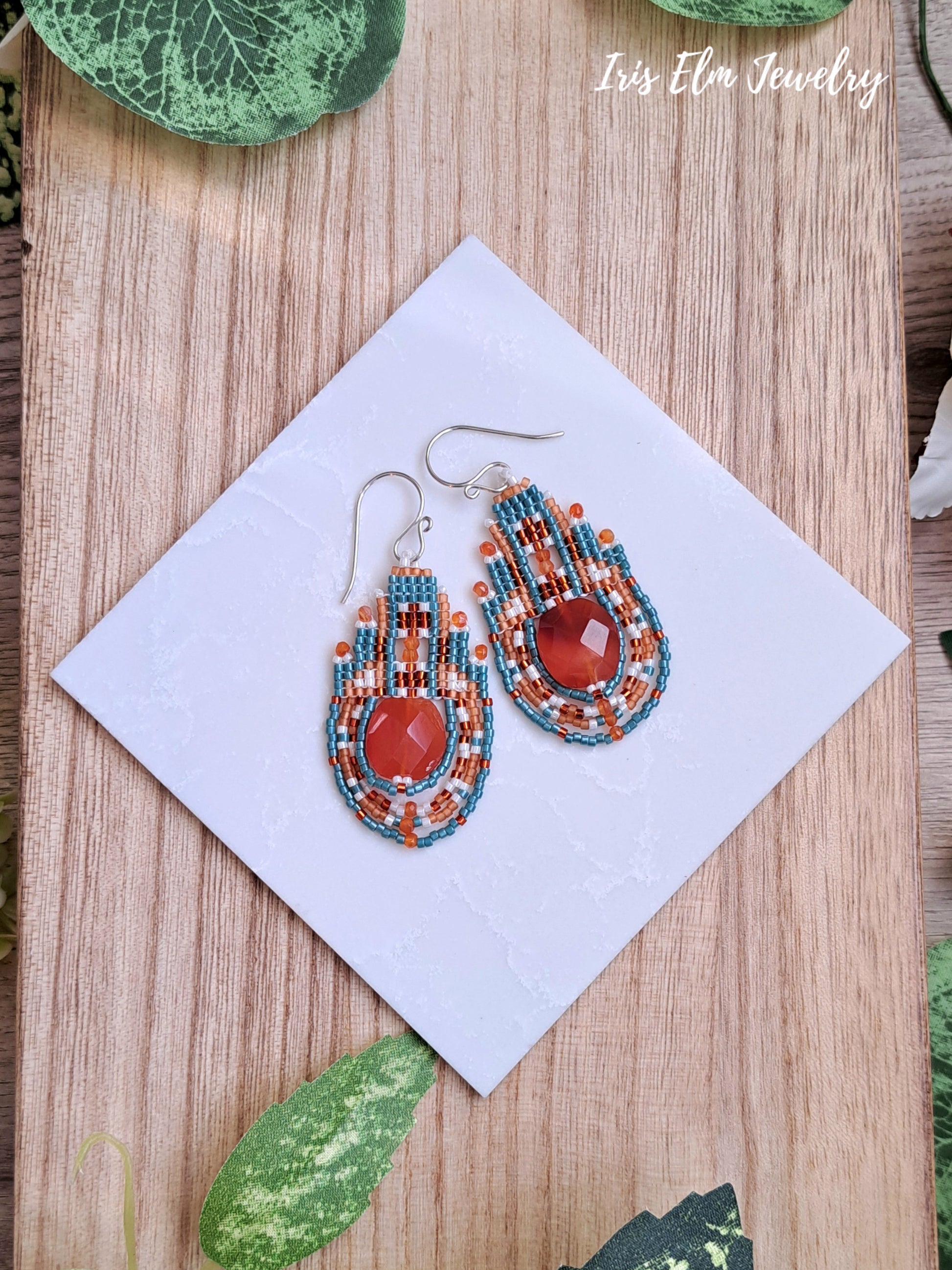Colorful beaded carnelian stone and seed bead handmade earrings on a white square with 'Iris Elm Jewelry' branding, placed on a wooden surface.