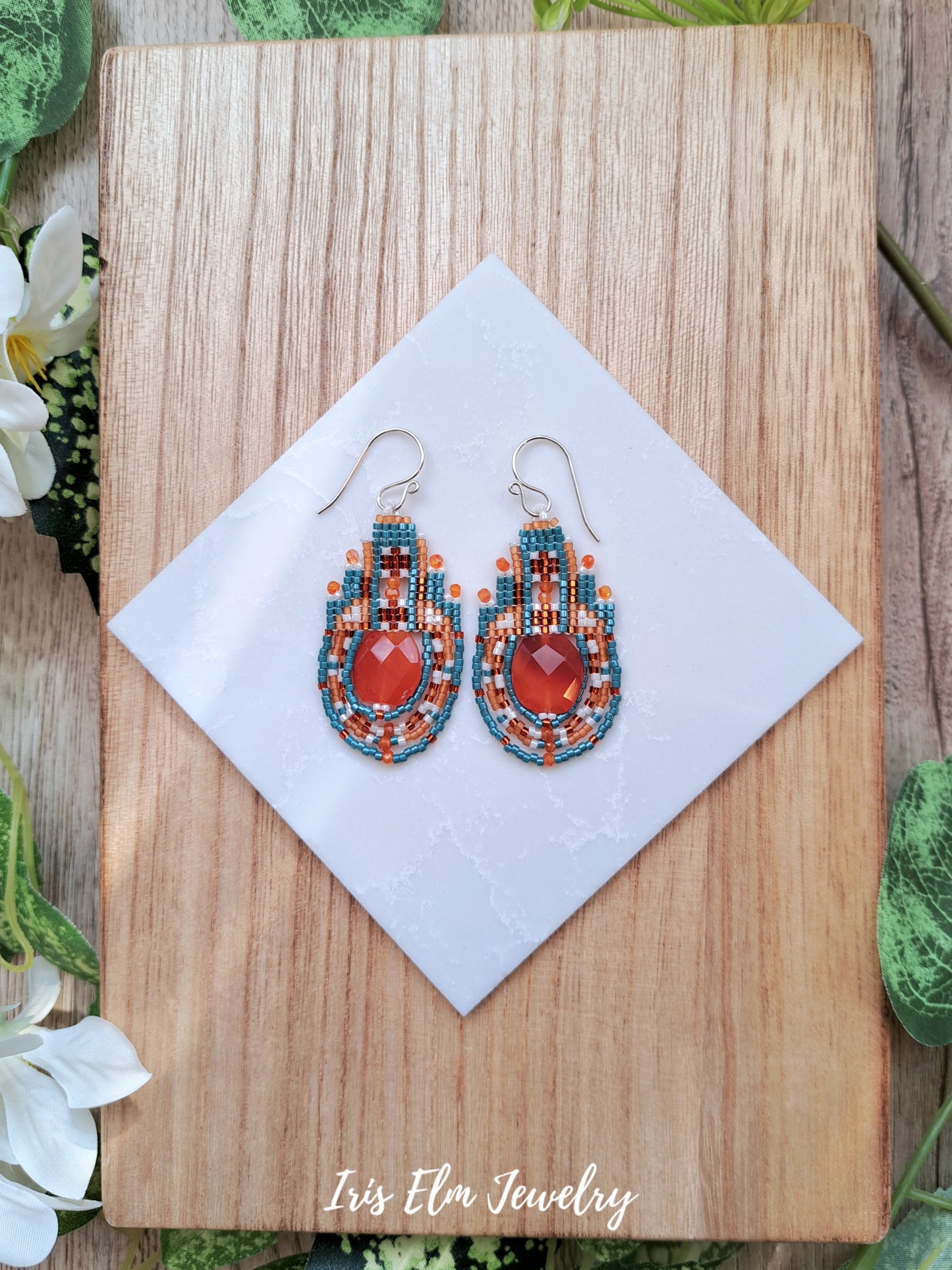 Pair of beaded carnelian earrings on a wooden board with a white tile, surrounded by green leaves and white flowers.