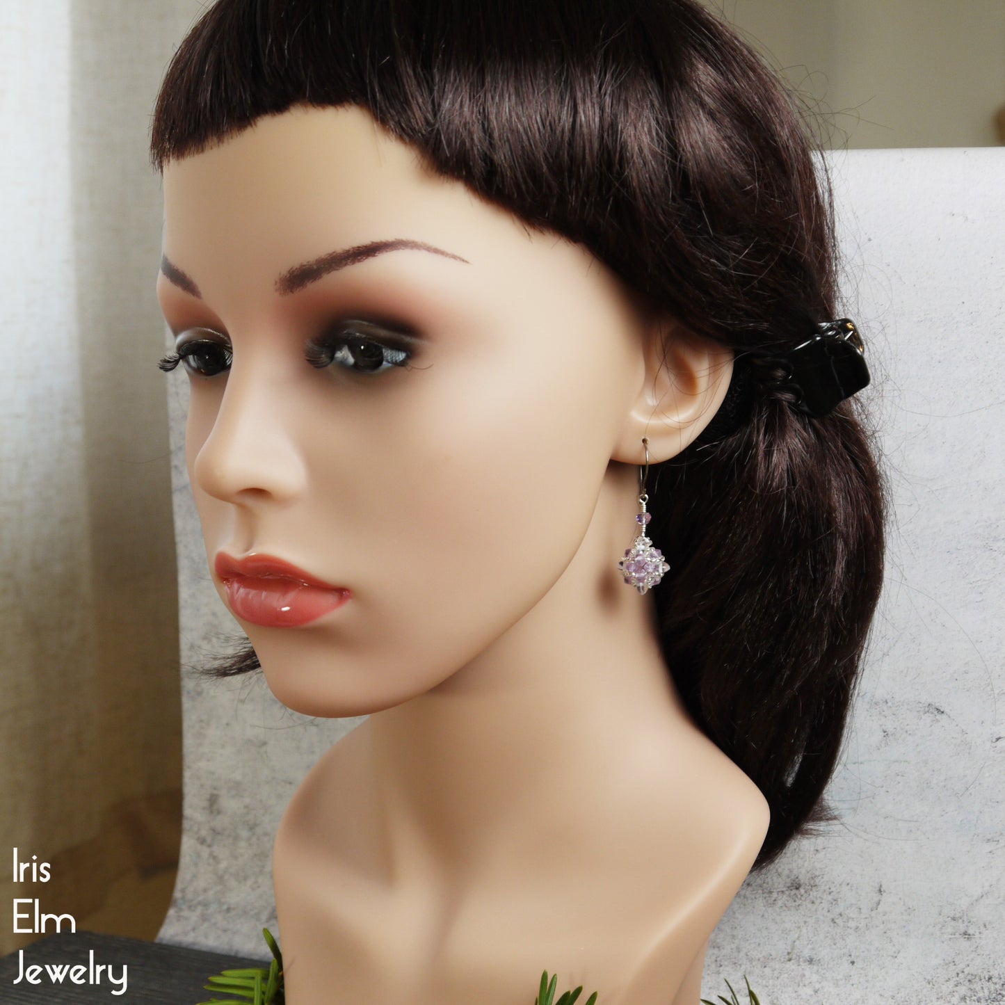 Mannequin head with dark hair and purple amethyst and clear quartz crystal earrings on a neutral background