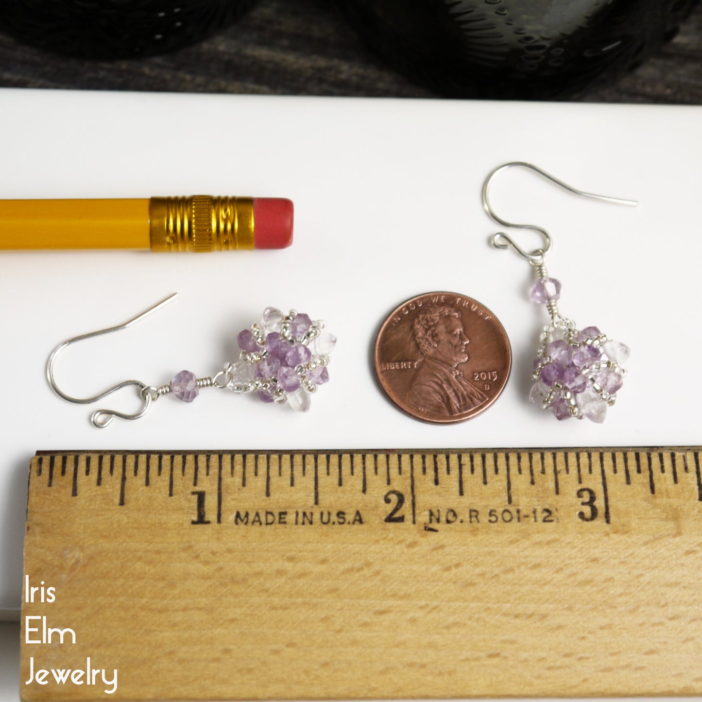Pair of 1.5 inch long silver earrings with purple beads on a white surface with a penny and ruler for scale, featuring 'Iris Elm Jewelry'.