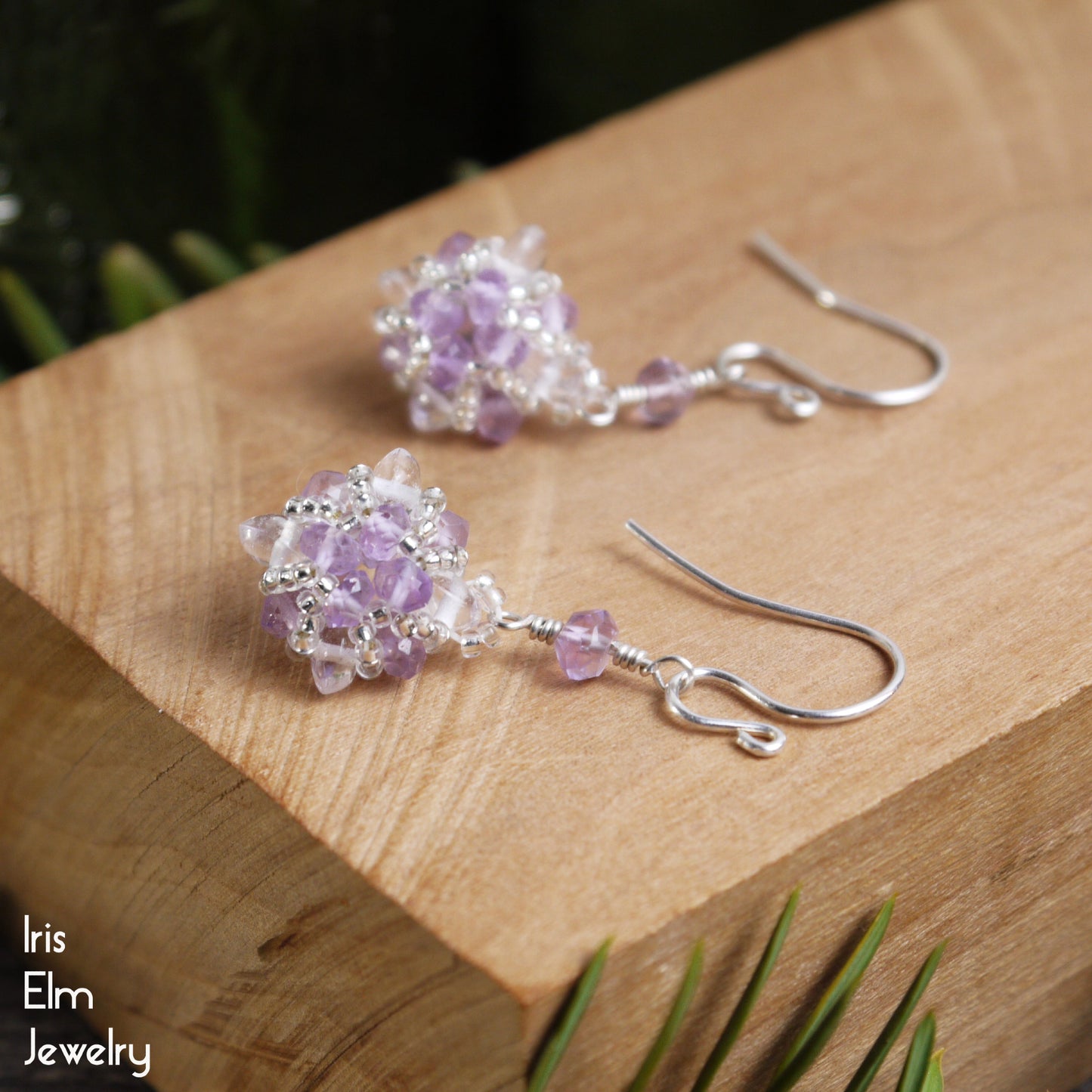 Pair of handmade sterling silver beaded purple amethyst and clear quartz crystal earrings on a wooden surface with 'Iris Elm Jewelry' branding.