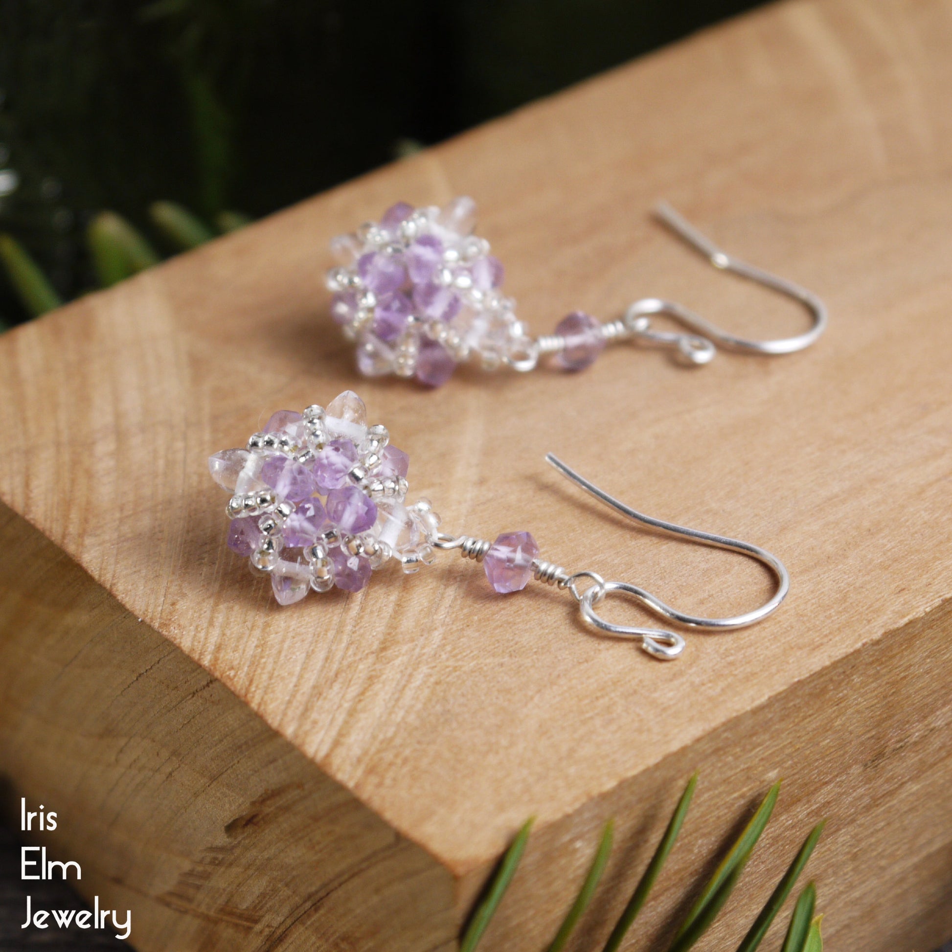 Pair of handmade sterling silver beaded purple amethyst and clear quartz crystal earrings on a wooden surface with 'Iris Elm Jewelry' branding.