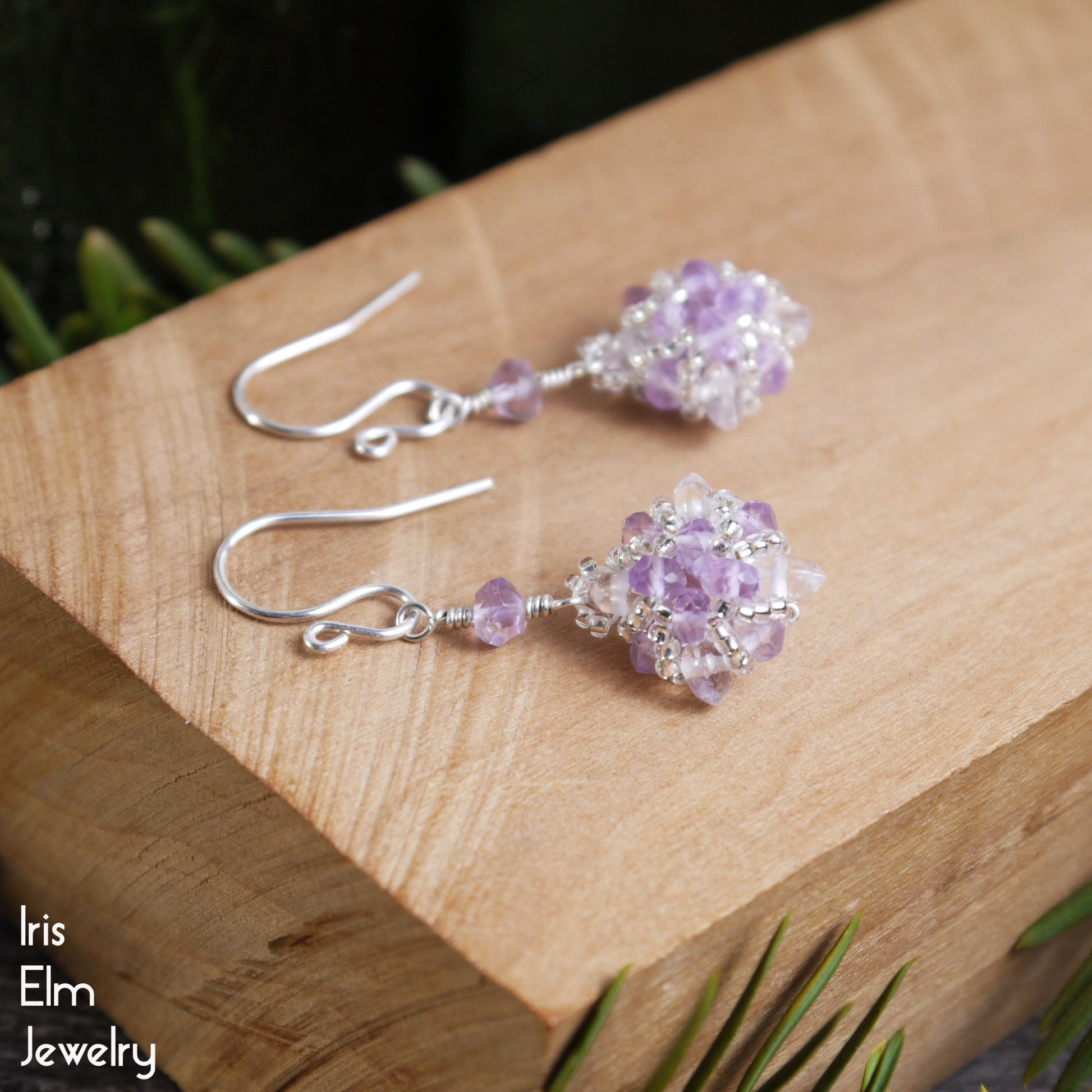 Pair of purple amethyst and clear quartz crystal beaded earrings on a wooden surface with 'Iris Elm Jewelry' branding.