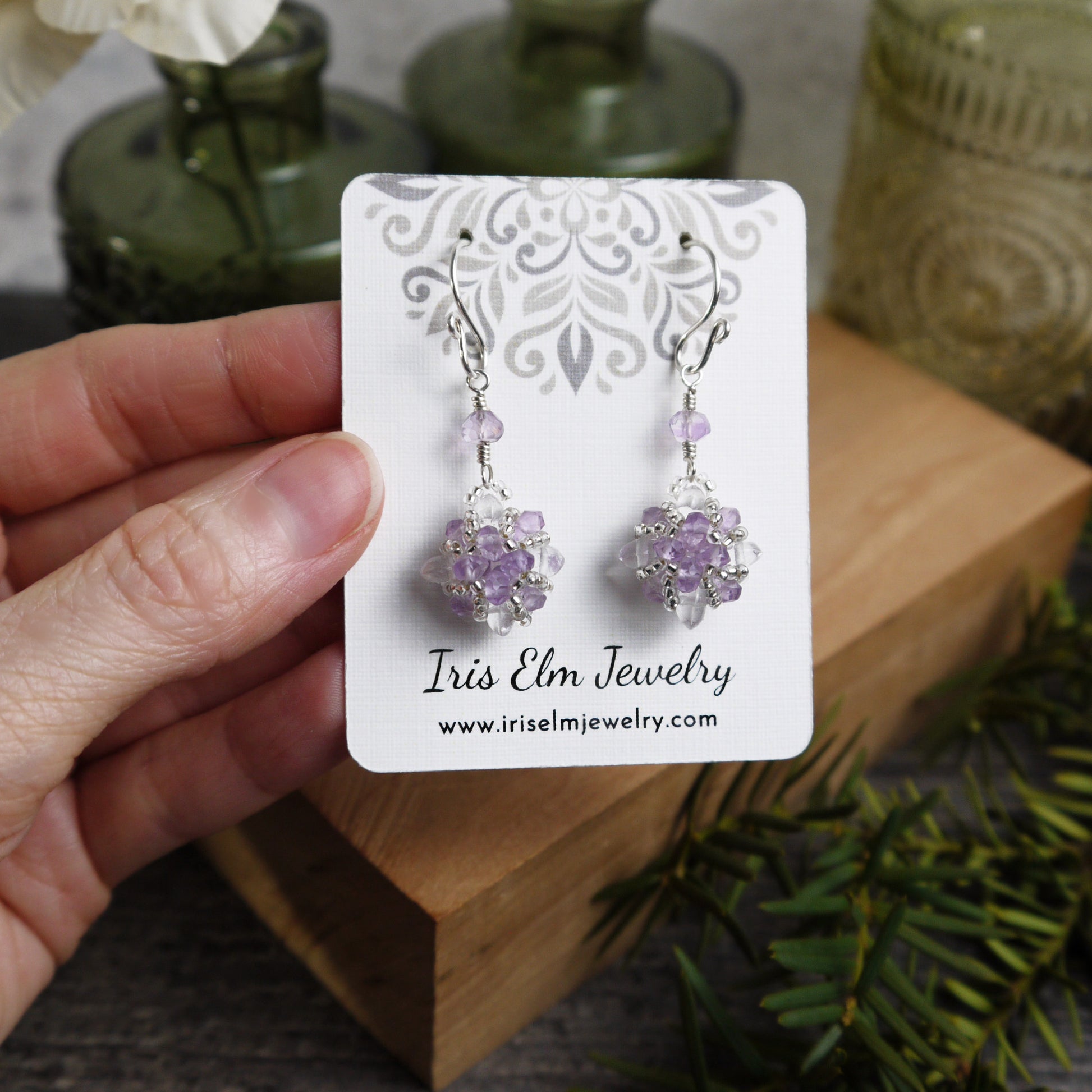 Hand holding a card with purple amethyst and clear quartz crystal earrings and 'Iris Elm Jewelry' branding.