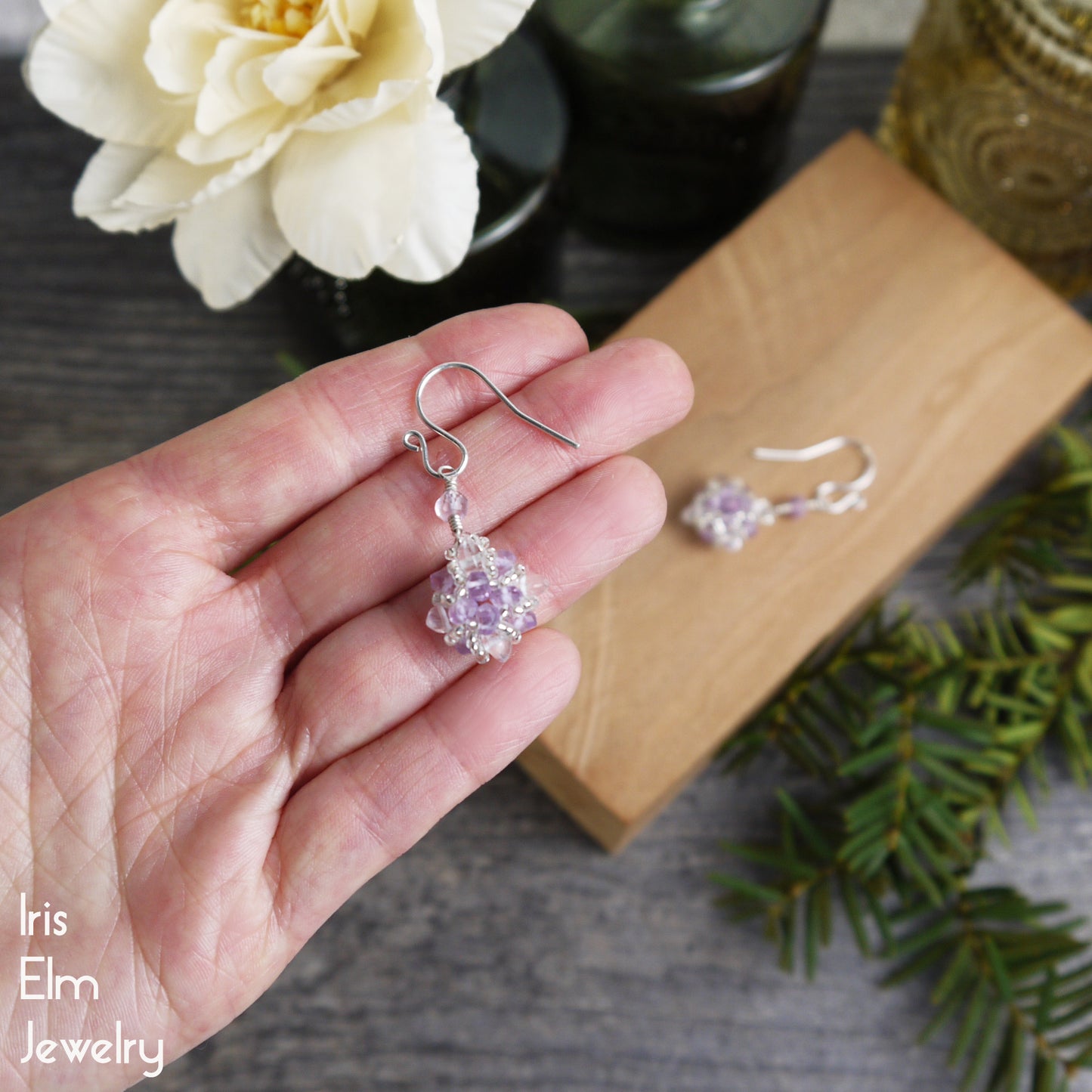 Hand holding a silver earring with a purple gemstone, placed on a wooden box with more earrings and decorative elements.