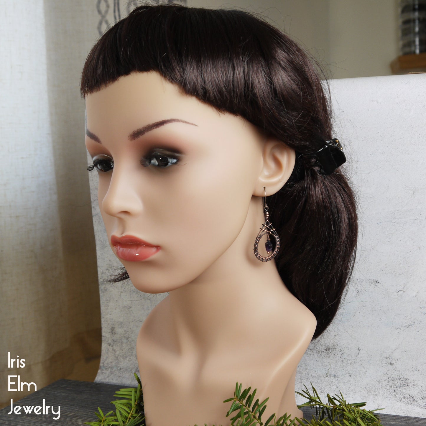 Mannequin head with dark hair styled in a ponytail, wearing copper amethyst earrings, with 'Iris Elm Jewelry' branding.