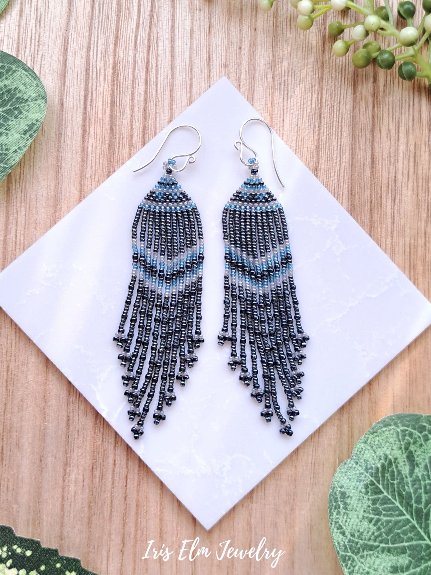 charcoal grey glass seed bead fringe handmade earrings on white tile surround by greenery with Iris Elm Jewelry branding