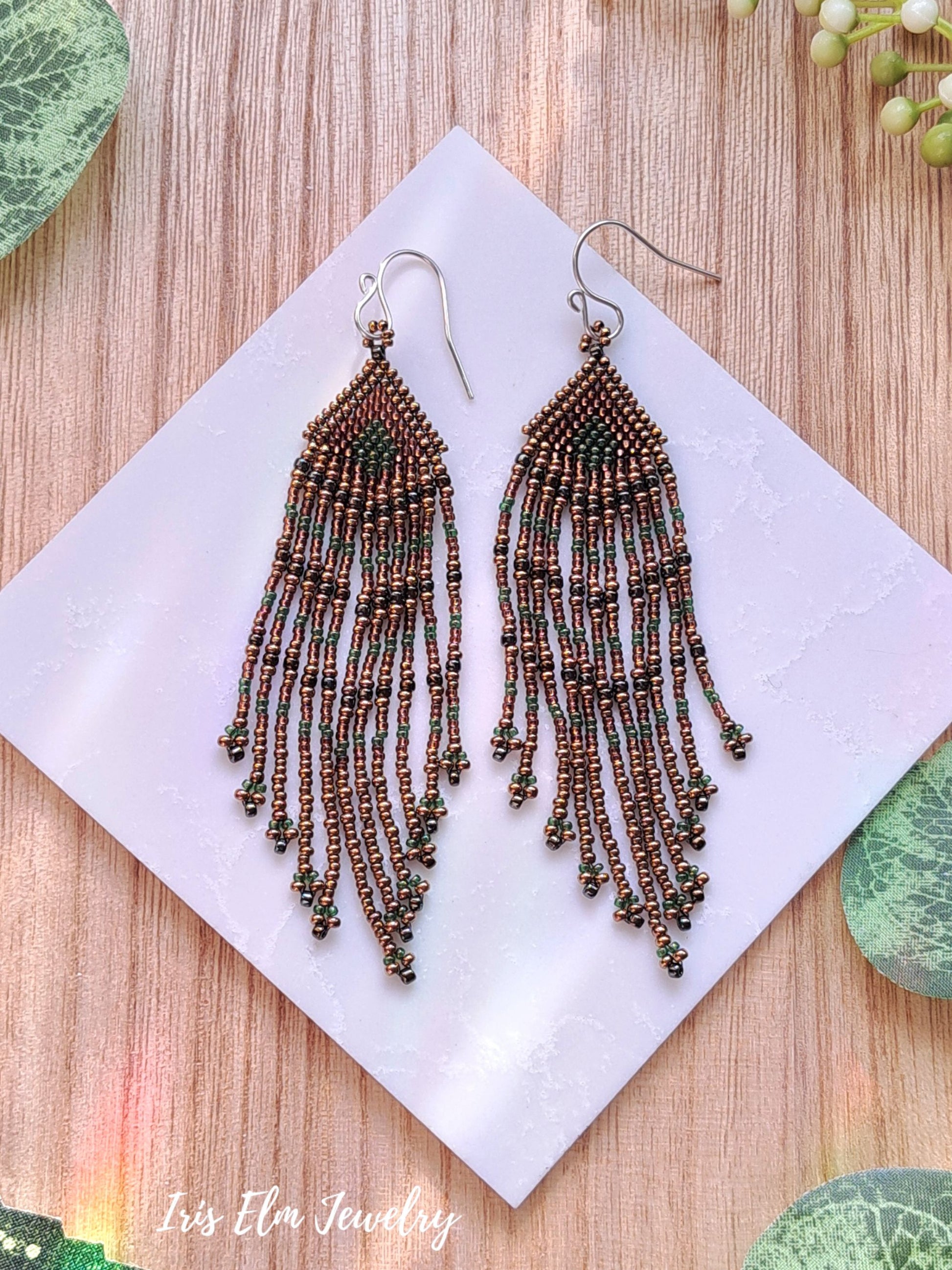 Beaded fringe bronze green brown seed bead fringe earrings on a white square with 'Iris Elm Jewelry' branding, placed on a wooden surface.