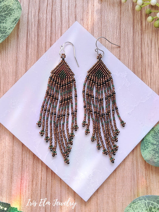 Beaded fringe bronze green brown seed bead fringe earrings on a white square with 'Iris Elm Jewelry' branding, placed on a wooden surface.