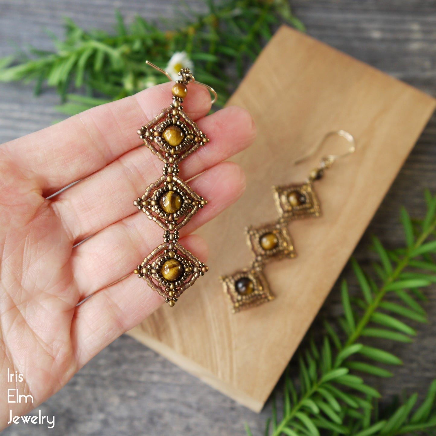 bronze seed bead and brown tiger eye beadwork earrings held by a hand with a wooden board and greenery in the background.