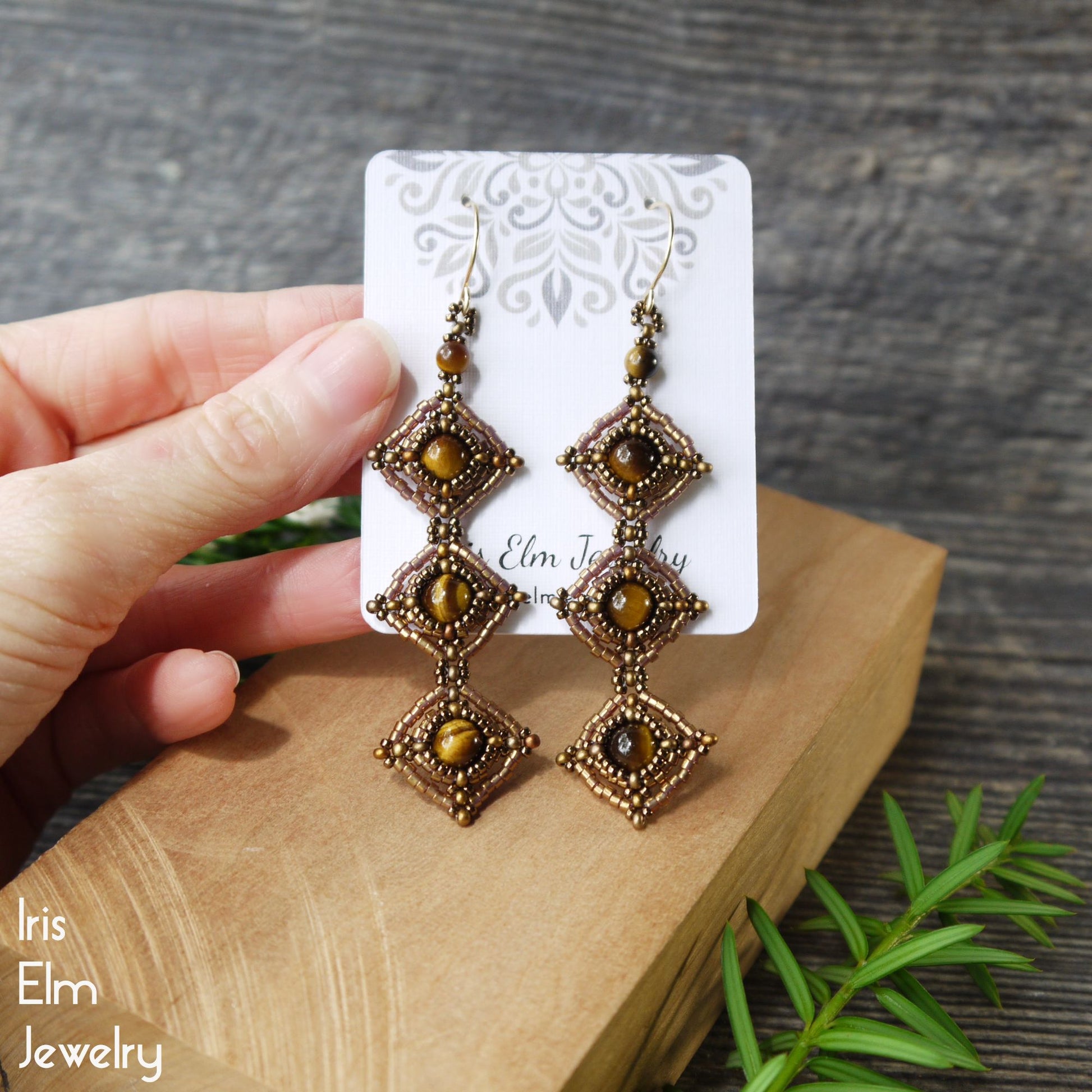 Pair of ornate tiger eye beaded earrings on a card held by a hand, with a wooden surface and greenery in the background.