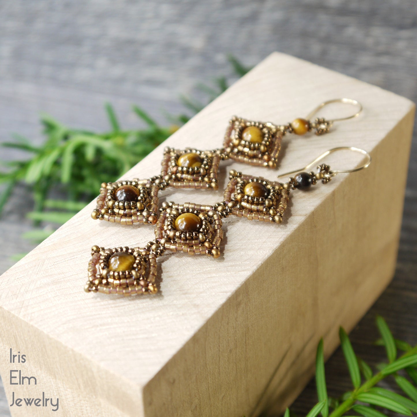 long beadwork earrings featuring a trio of brown tiger eye stones surrounded by bronze glass seed beads on a wooden block with greenery in the background, featuring the brand 'Iris Elm Jewelry'.