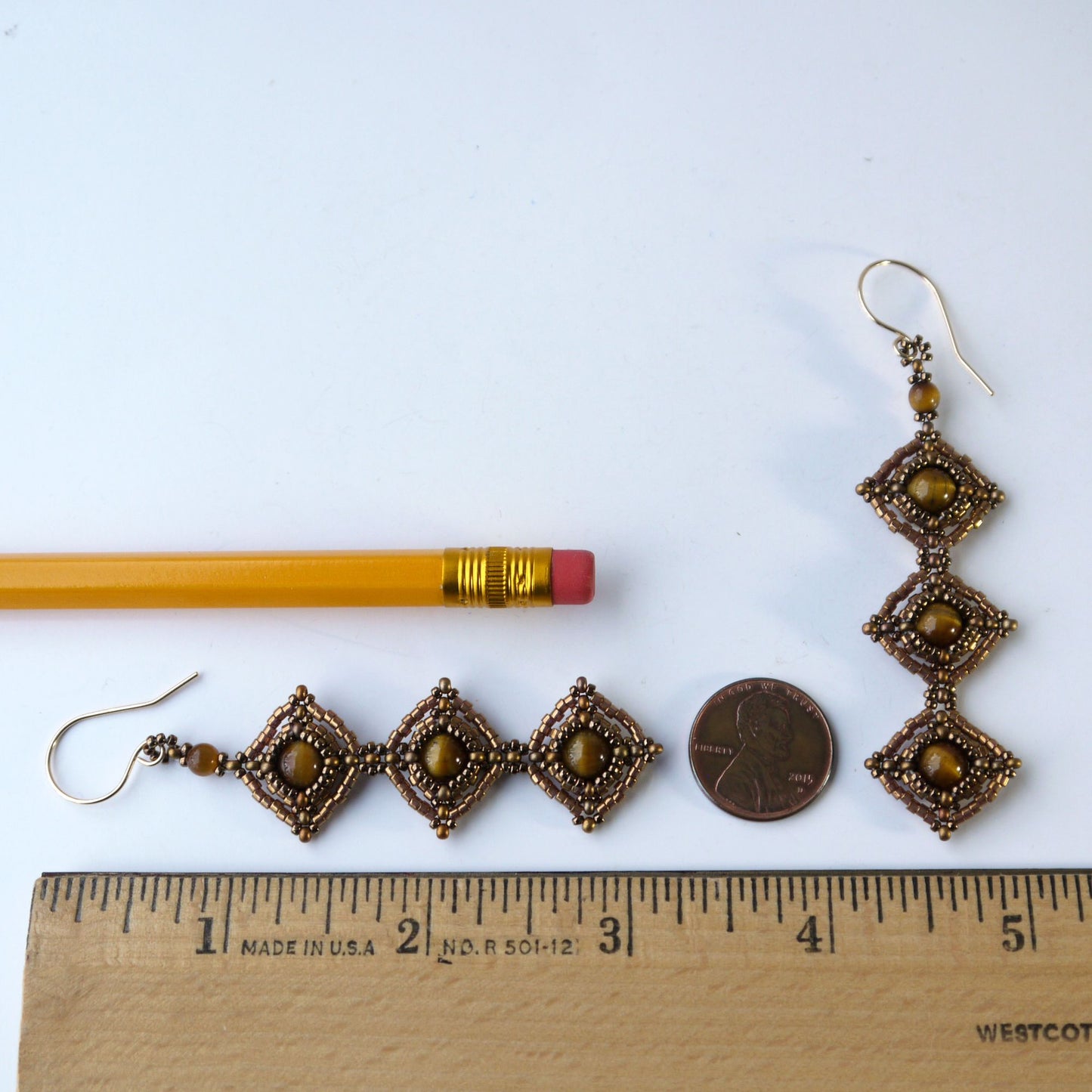Pair of decorative 3.25 inch long earrings with brown tiger eye stones, a pencil, and a coin for scale on a white background.