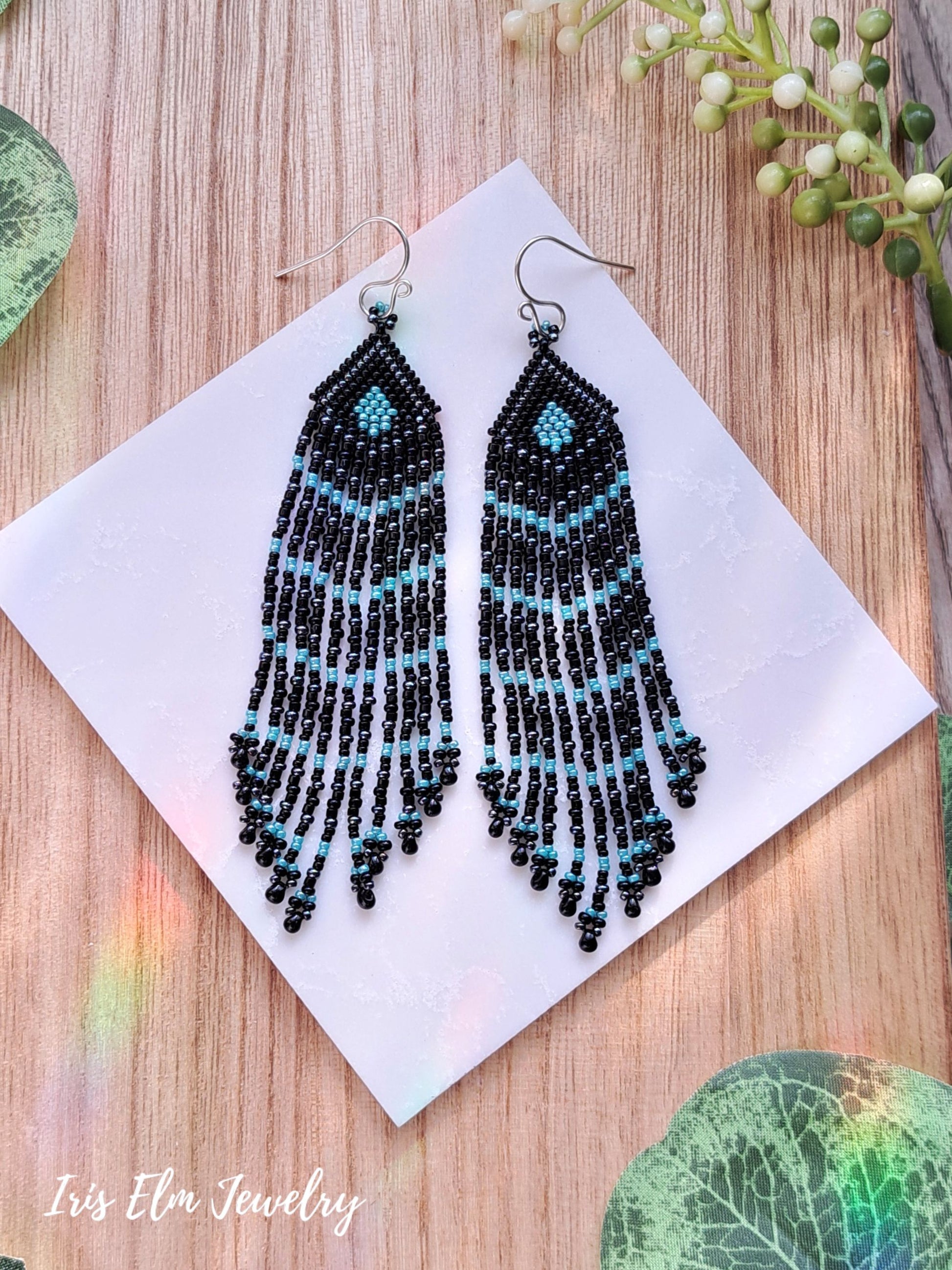 Black and turquoise beaded seed bead fringe earrings on a white card with a wooden background