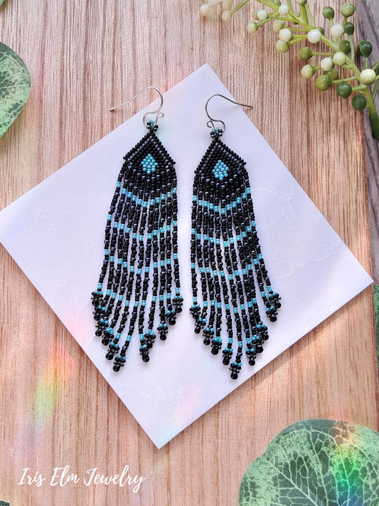 Black and turquoise beaded seed bead fringe earrings on a white card with a wooden background