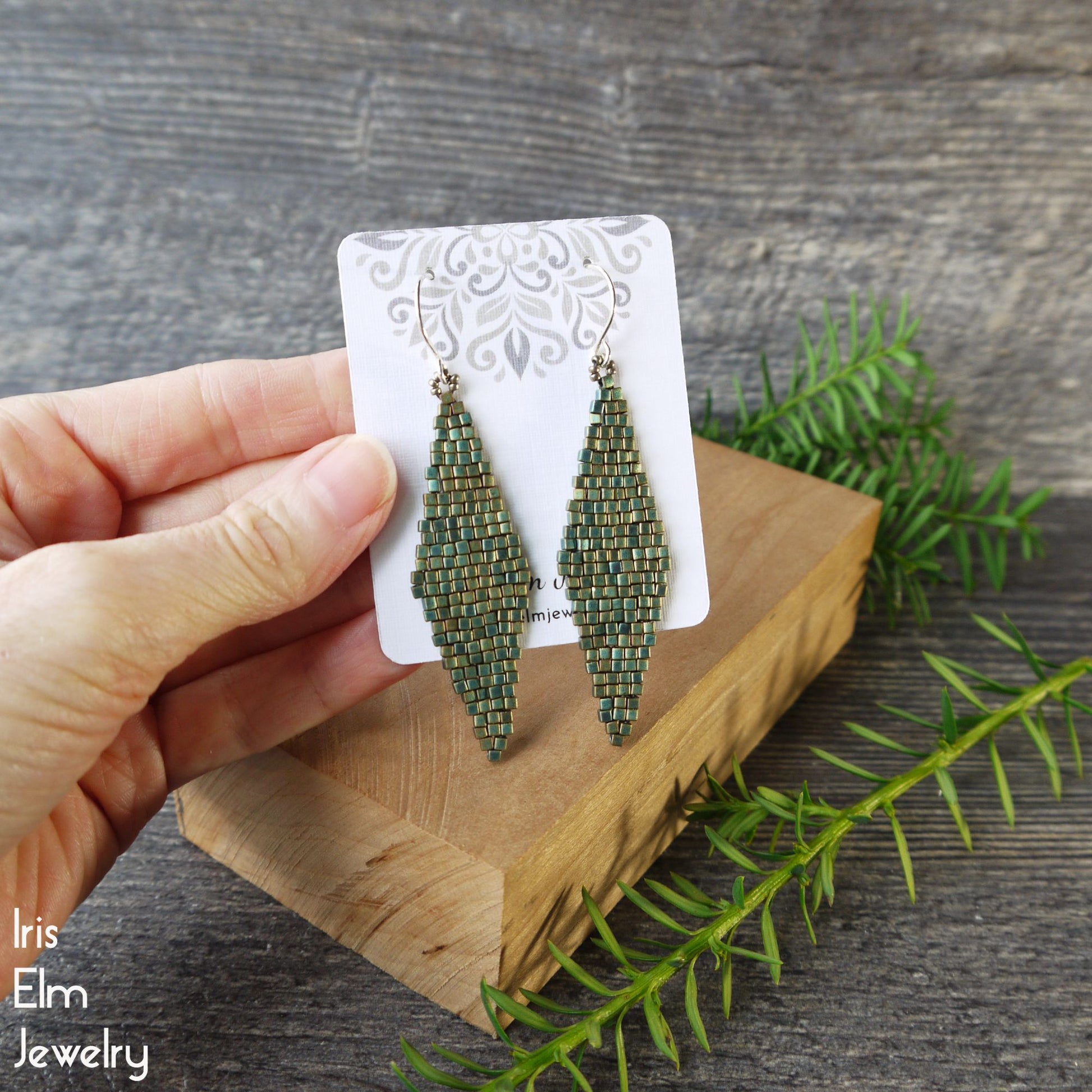 turquoise teal Green beaded earrings on a card held by a hand with a wooden block and green leaves in the background.