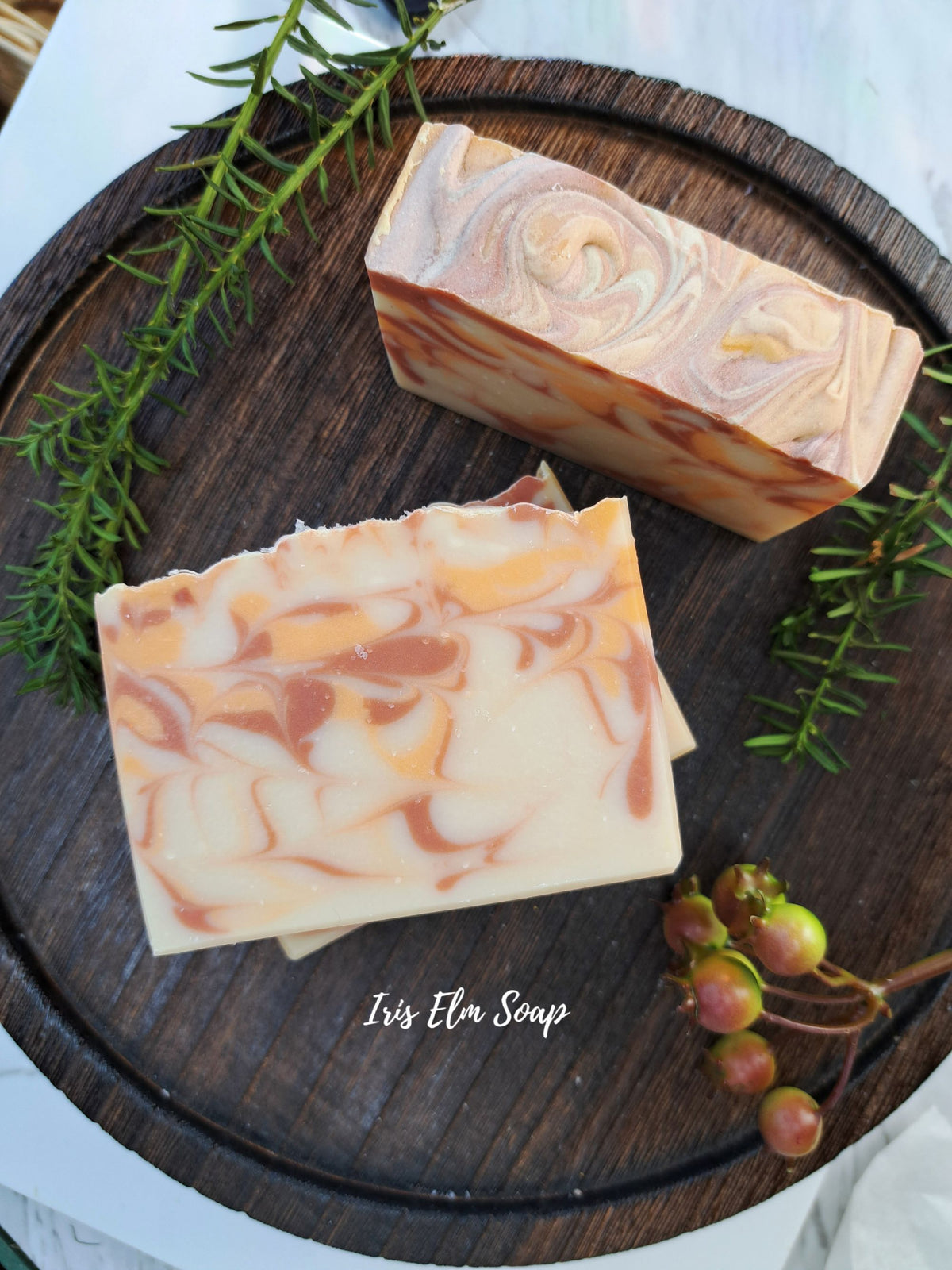 Two rectangular bars of swirled orange cranberry spice soap with peach, orange, and cream tones arranged on a dark round wooden tray, with green sprigs and small berries as styling props. The text 'Iris Elm Soap' appears near the bottom.