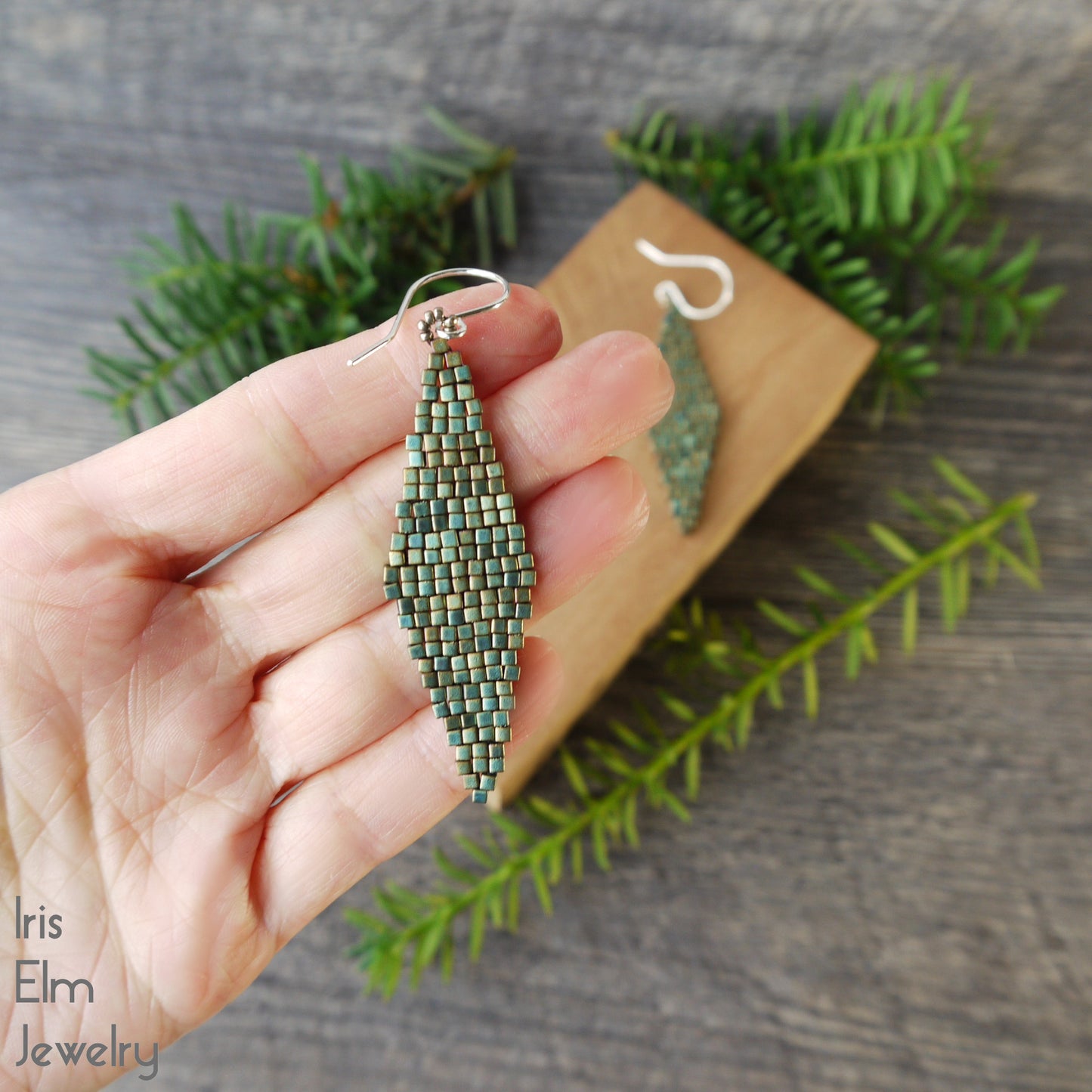 Hand holding a turquoise green beaded diamond shape woven matte glass bead earring with 'Iris Elm Jewelry' branding on a wooden block and green leaves.