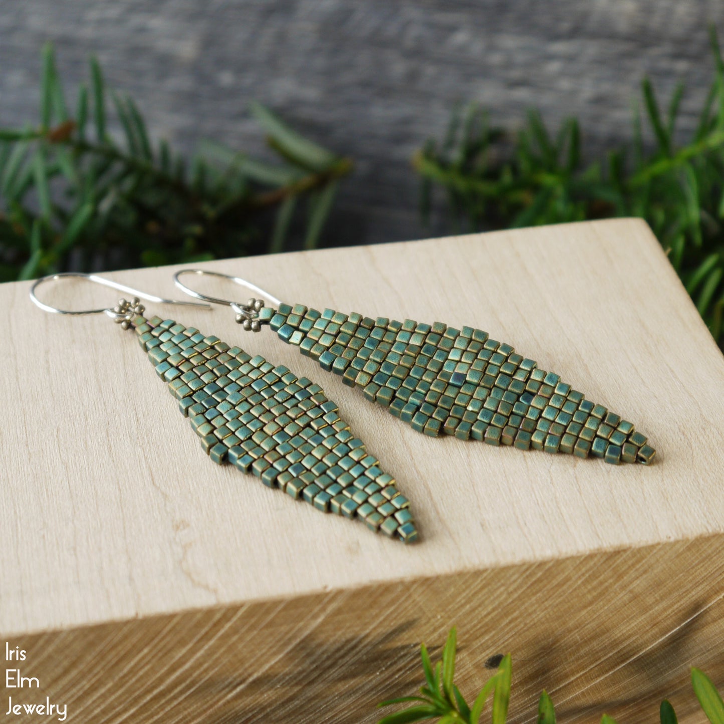 Teal beaded diamond shape earrings on a wooden block with greenery in the background