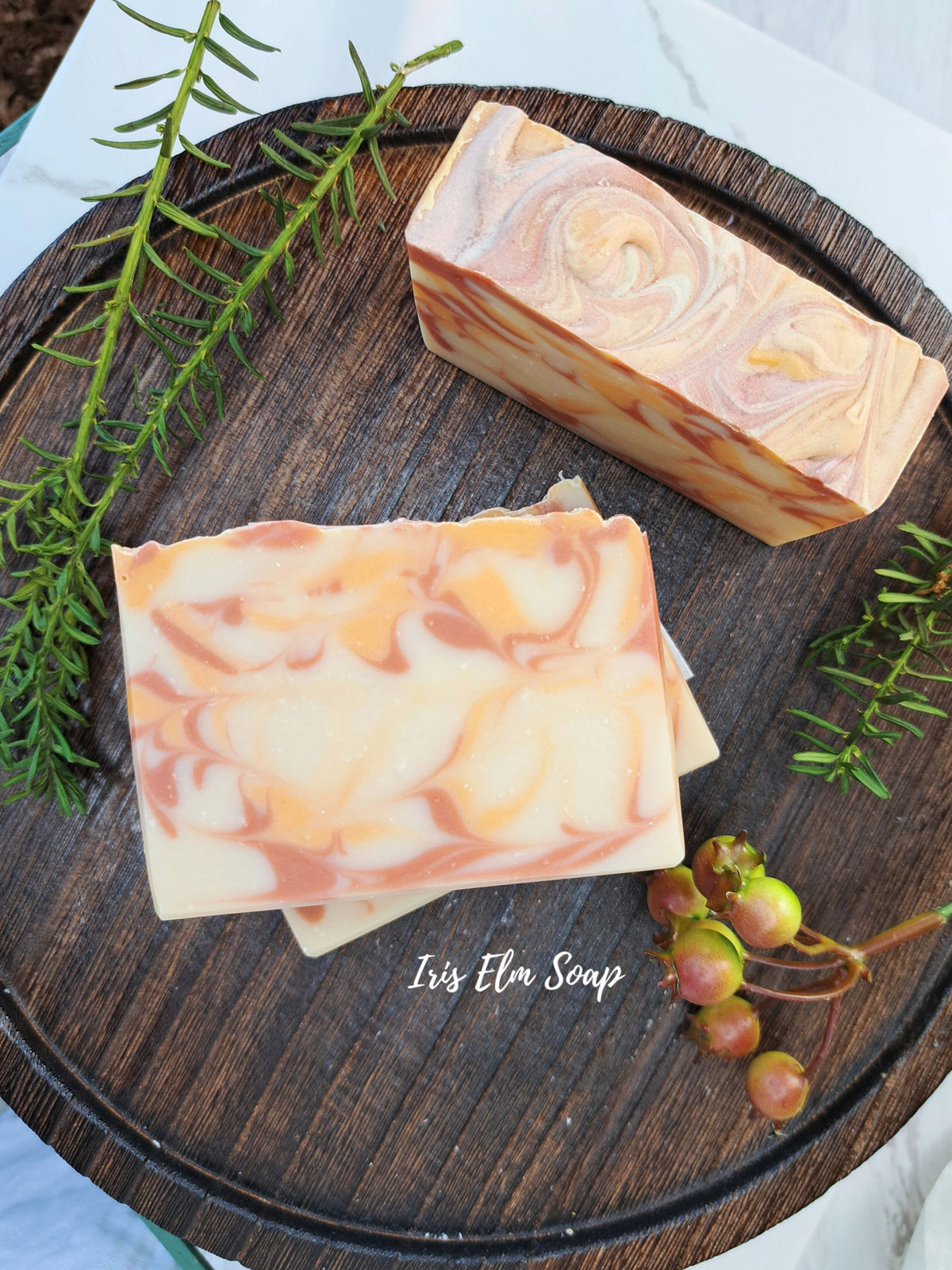 Two rectangular bars of marbled orange cranberry spice artisan soap in peach, cream, and orange tones, arranged on a dark wooden tray with green sprigs and small berries. The soap bar in the foreground is stacked on a second bar, and a second bar sits upright in the background.