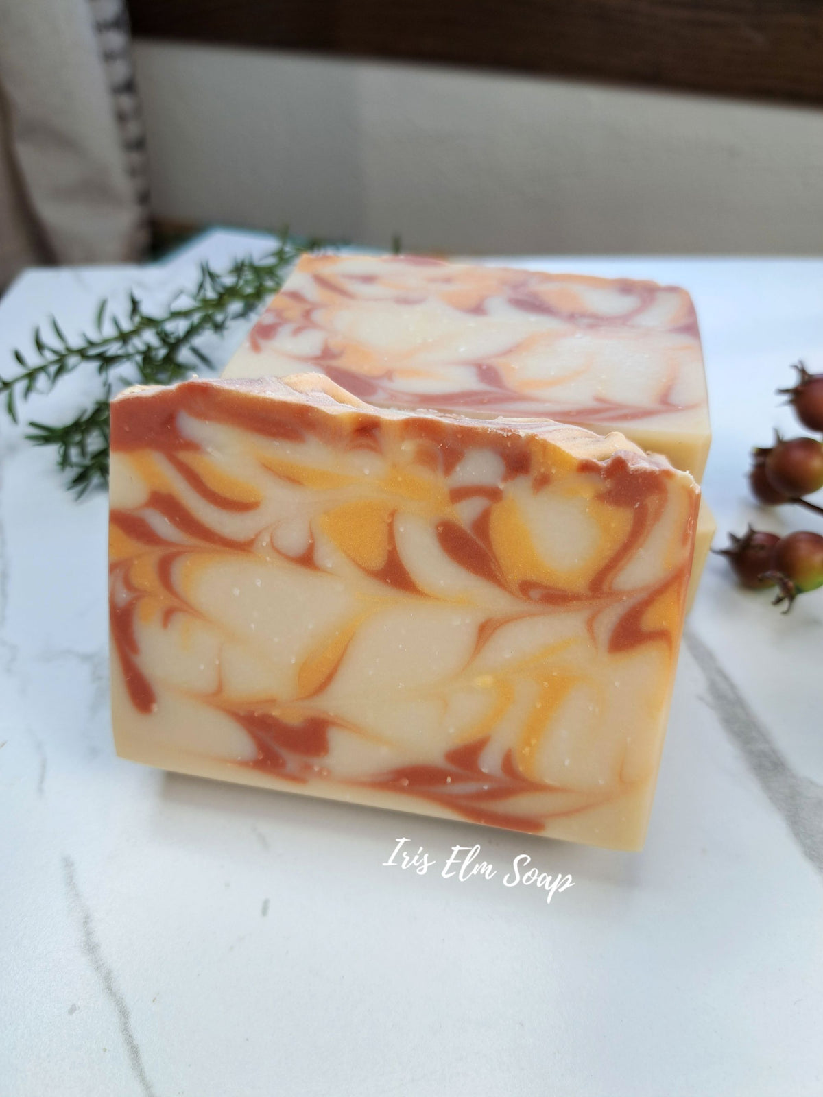 Two rectangular bars of artisanal orange cranberry spice soap with a marbled swirl pattern of cream, orange, and brown, displayed on a white marble surface with rosemary and small rosebuds as decorative props. The brand name 'Iris Elm Soap' is written in white script on the front bar.