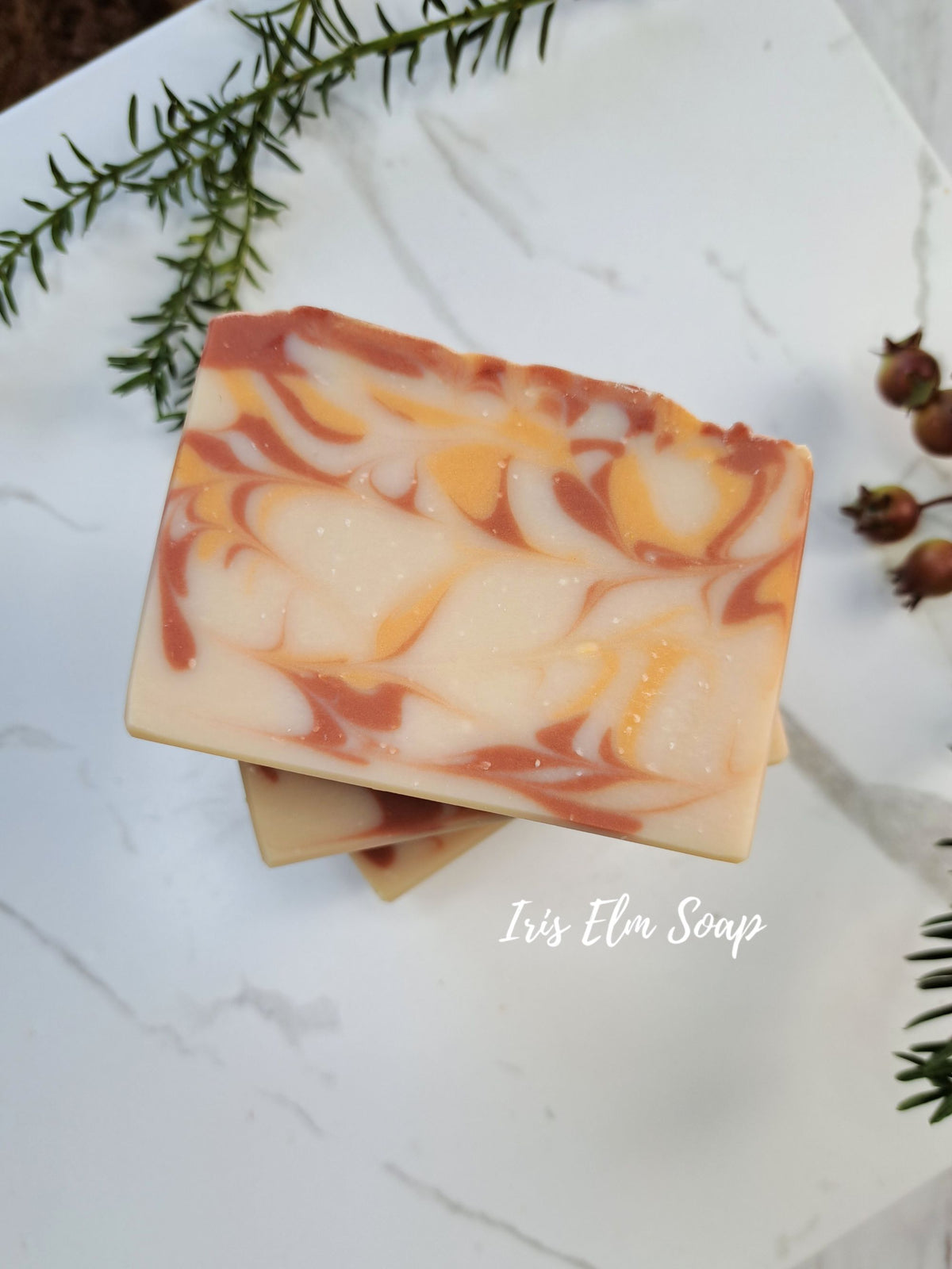 A handmade rectangular bar of soap with a marbled swirl pattern of cream, orange, and terracotta brown, stacked on another bar on a white marble surface with green sprigs and small dried flowers as props. The text 'Iris Elm Soap' is visible in the lower right.