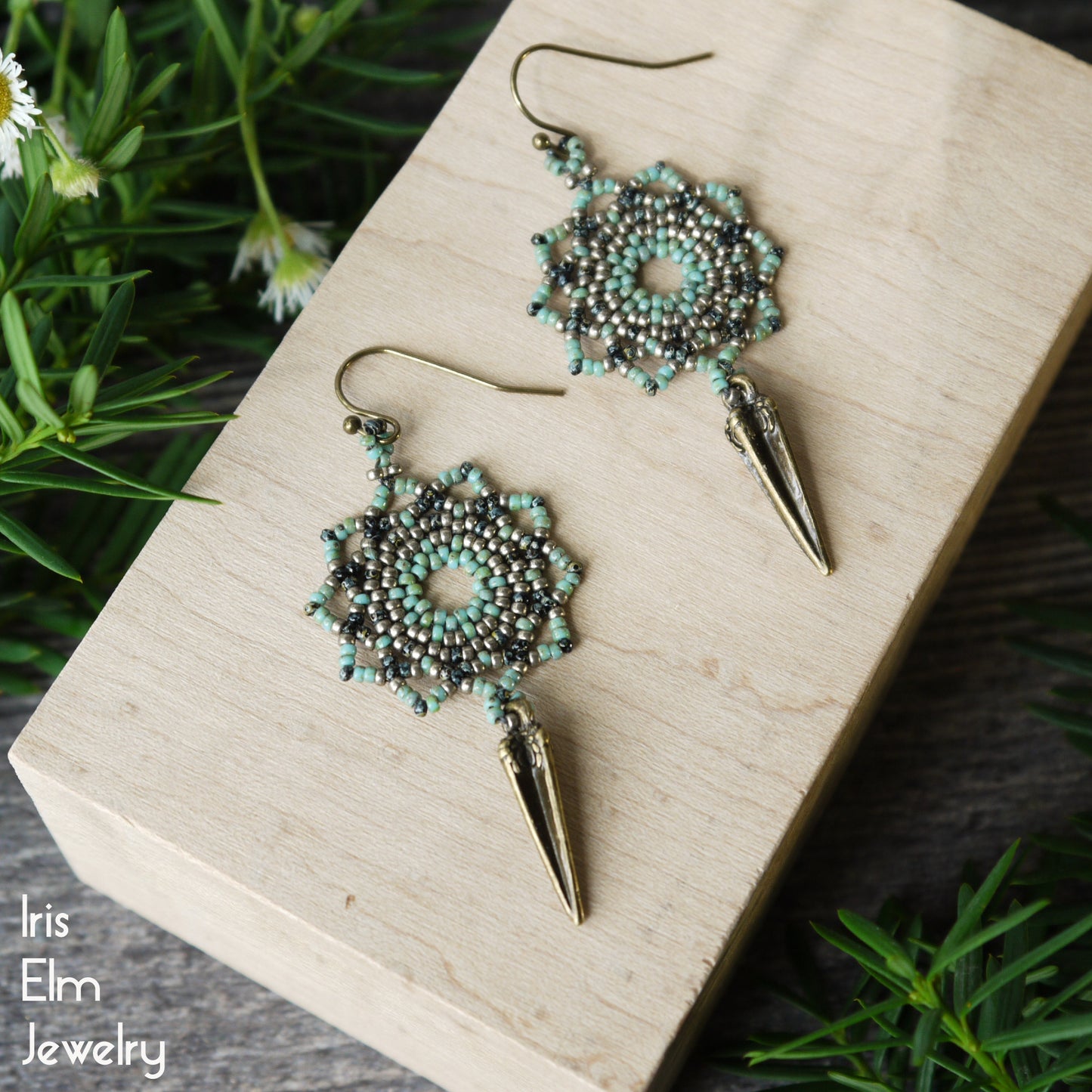 Pair of green seed bead woven mandala earrings with antiqued brass spike details on a wooden block, surrounded by greenery.