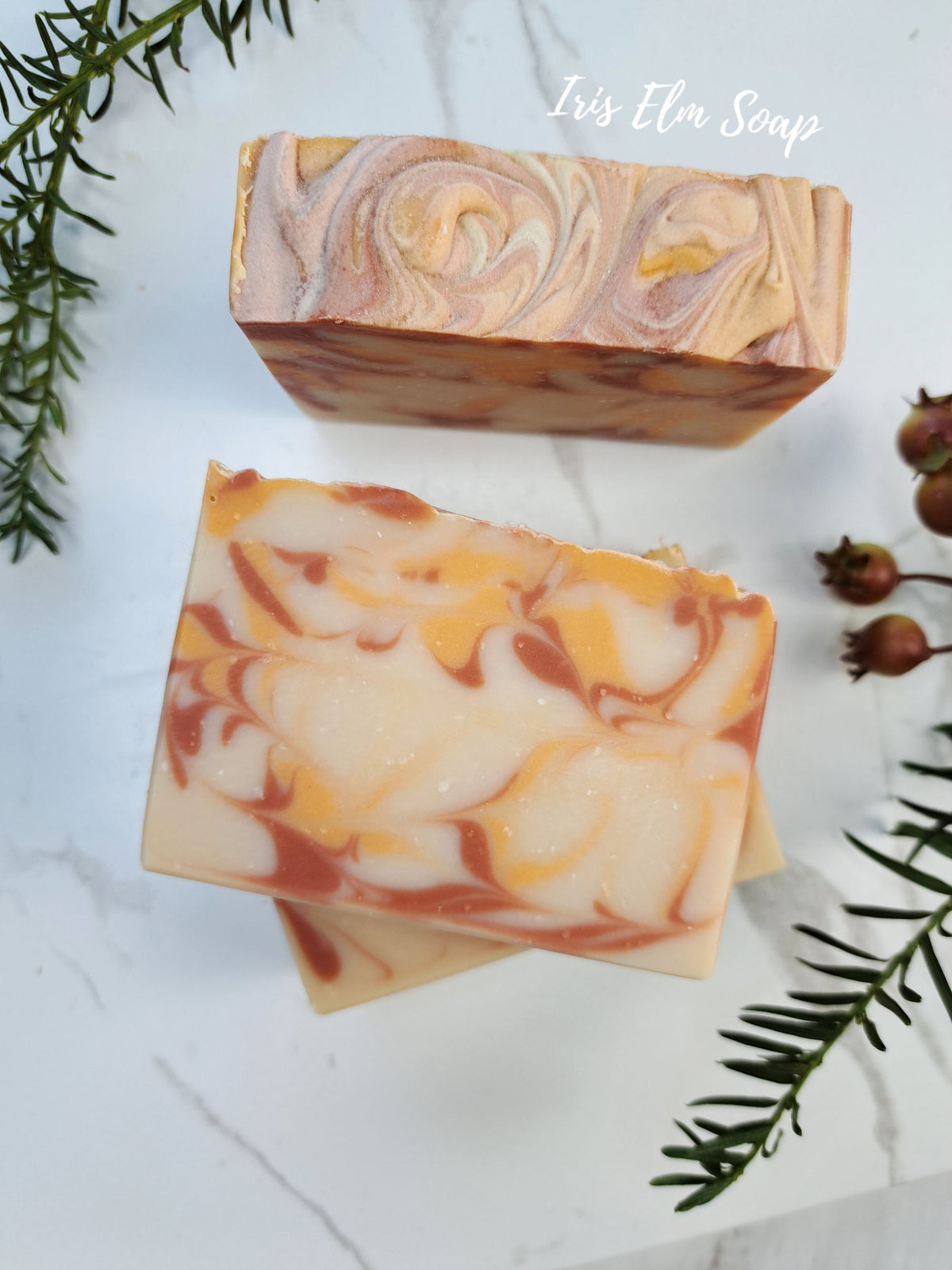 Two rectangular artisan orange cranberry spice soap bars with marbled swirls of cream, orange, and terracotta red, displayed on a white marble surface with green pine branches and small berries. The text 'Iris Elm Soap' is visible in the upper right.