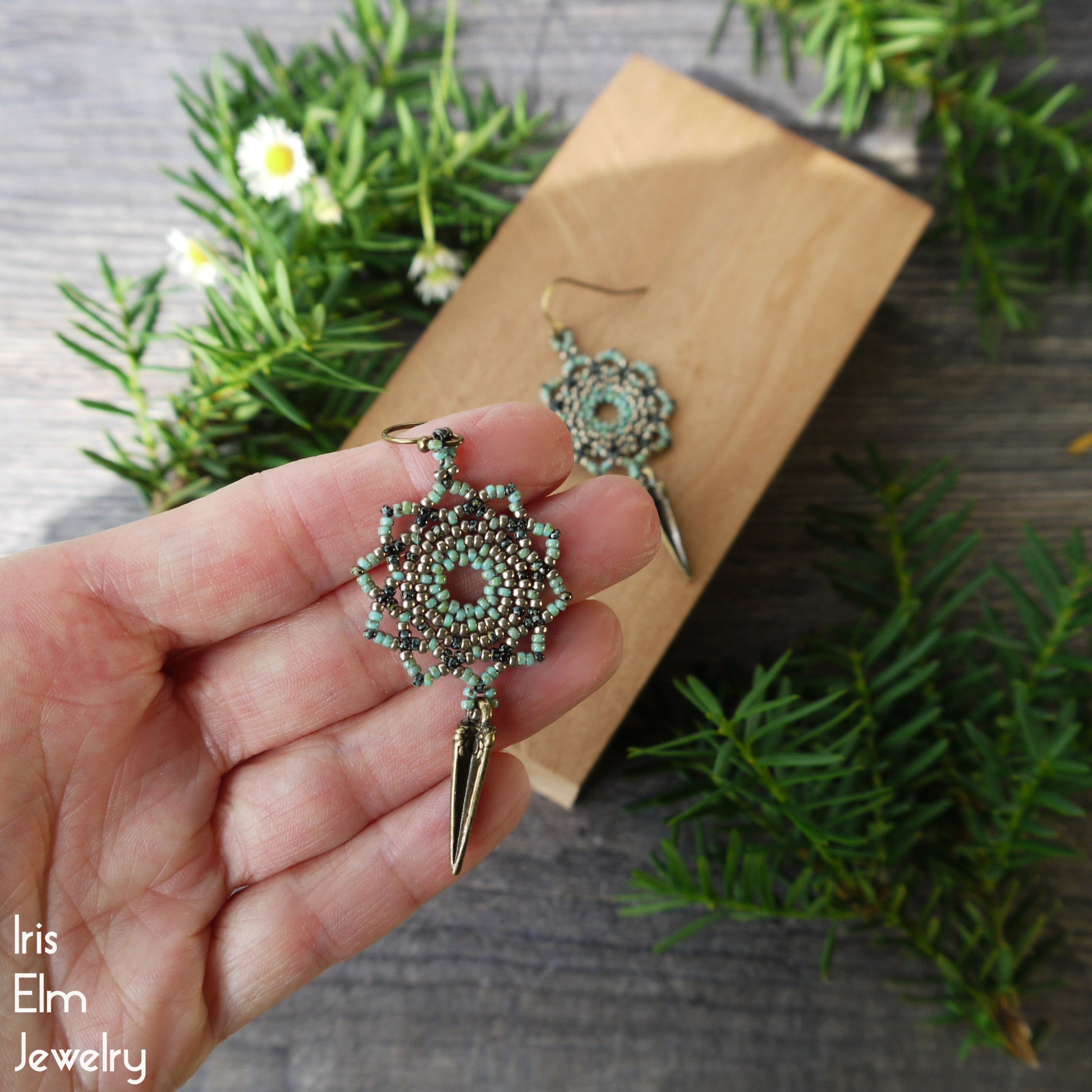 Hand holding a seafoam green mandala earring with plants in the background, featuring 'Iris Elm Jewelry'.