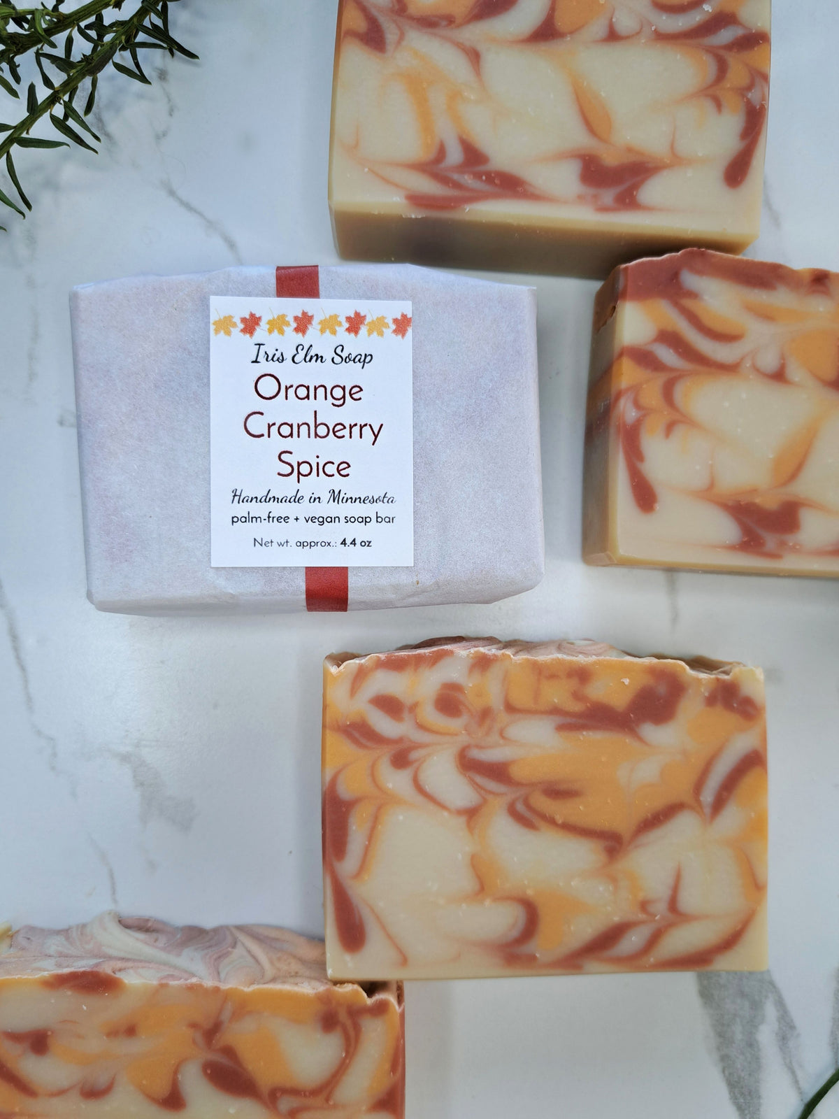 Several handmade soap bars are arranged on a light marble surface. One bar is wrapped in pale pink paper with a white label reading 'Iris Elm Soap Orange Cranberry Spice,' and the others show a marbled swirl of creamy off-white soap with orange and reddish-brown streaks.