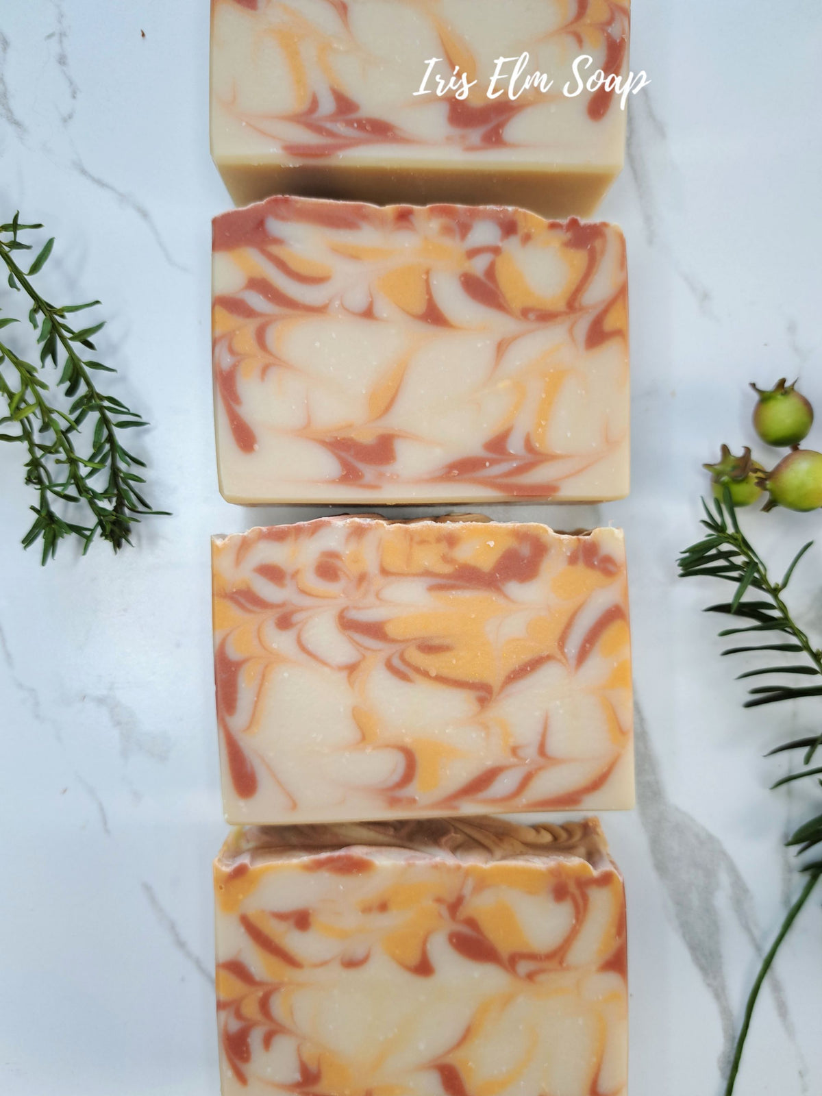 Four rectangular bars of artisanal orange cranberry spice soap stacked vertically on a white marble surface, decorated with green rosemary sprigs and small green berries. The soap has a marbled pattern of cream, orange, and reddish-brown swirls.