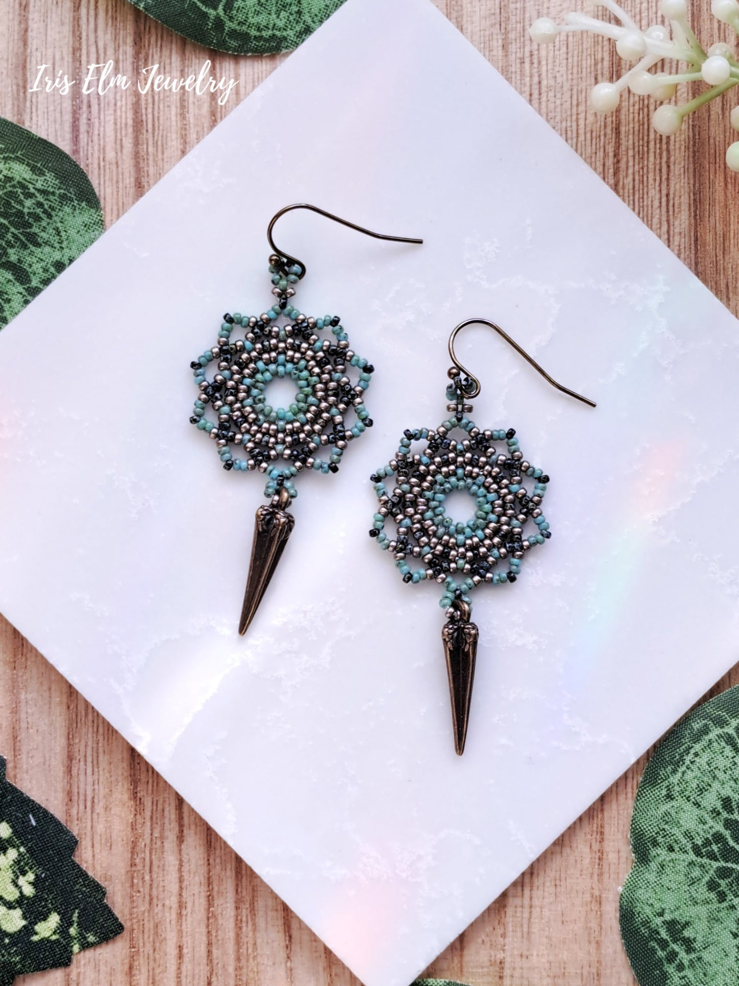 Pair of beaded mandala earrings with spike details on a white card with green leafy background