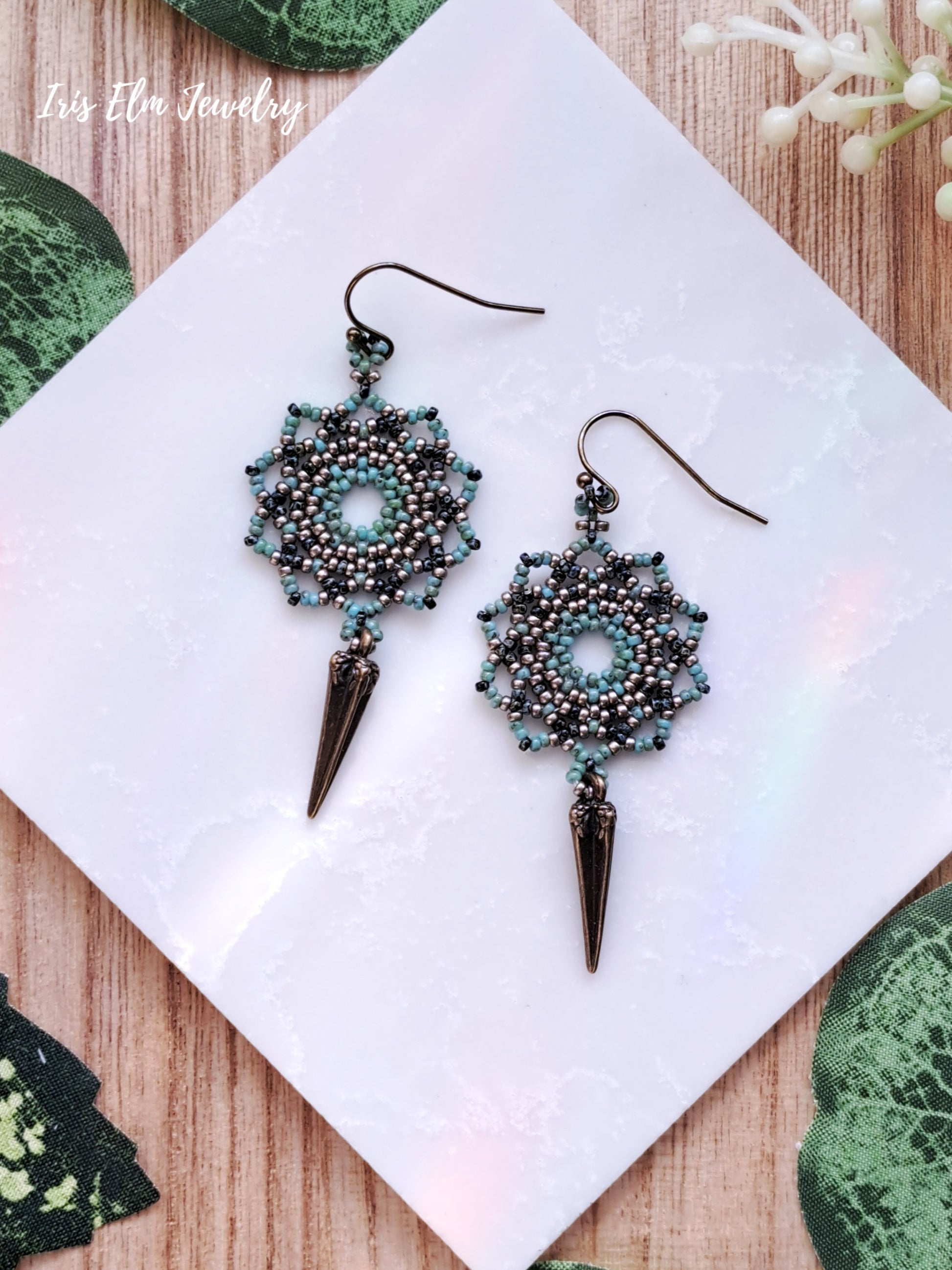 Pair of beaded mandala earrings with spike details on a white card with green leafy background