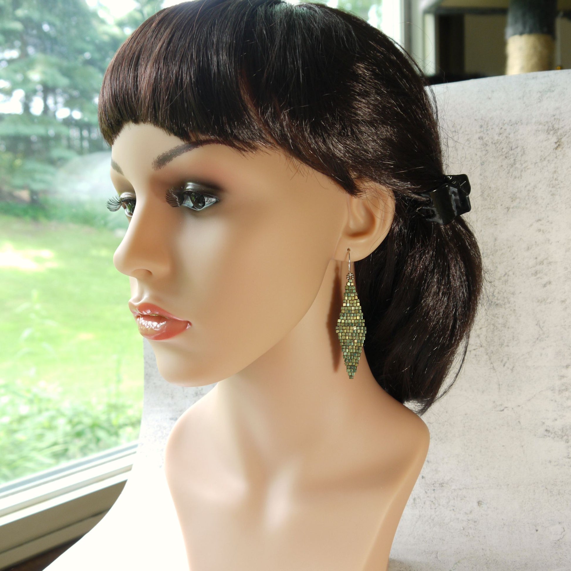 Mannequin head with dark hair and long turquoise green diamond shape glass earrings in front of a window with greenery.