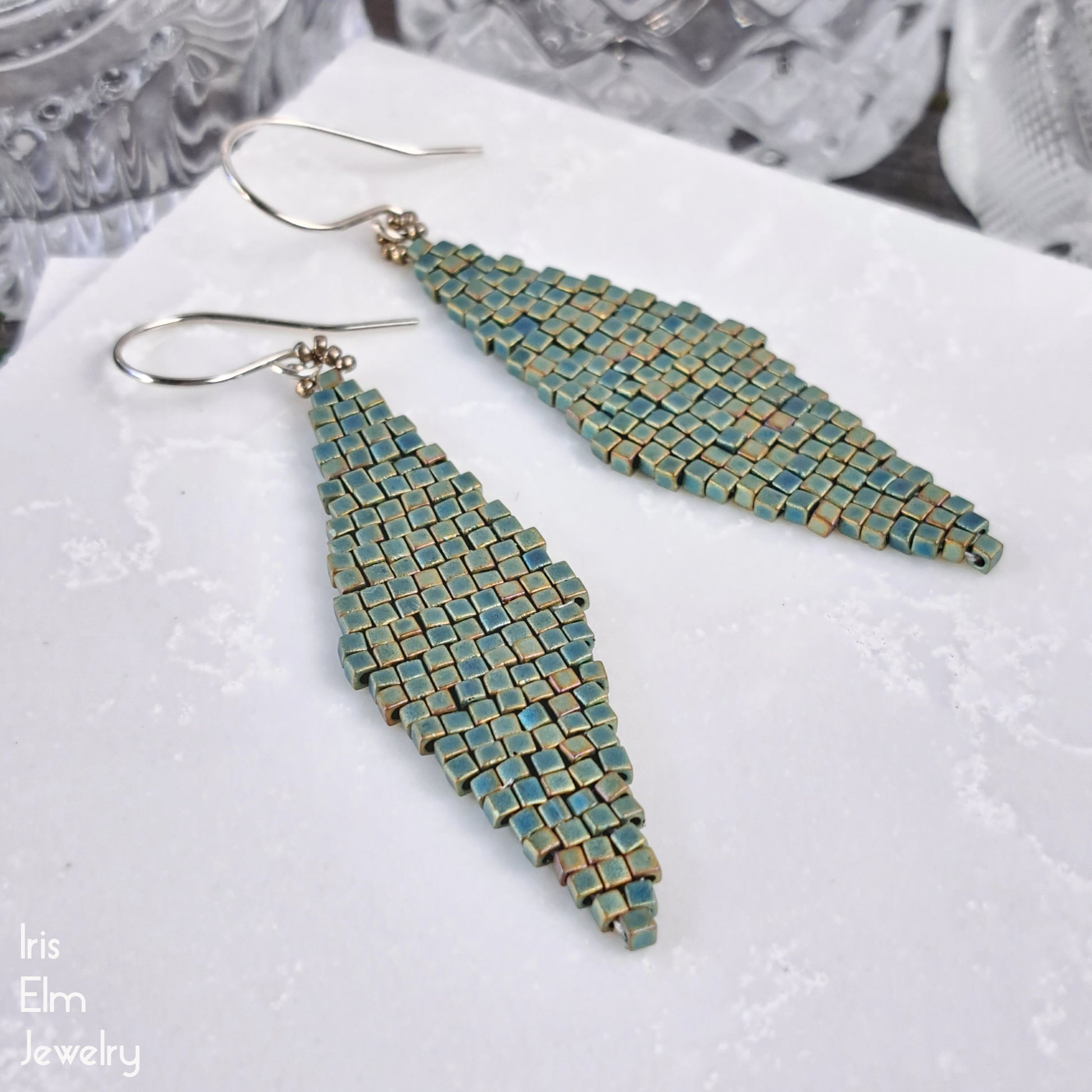 Pair of teal beaded diamond shape matte glass earrings on a textured white surface with 'Iris Elm Jewelry' branding.