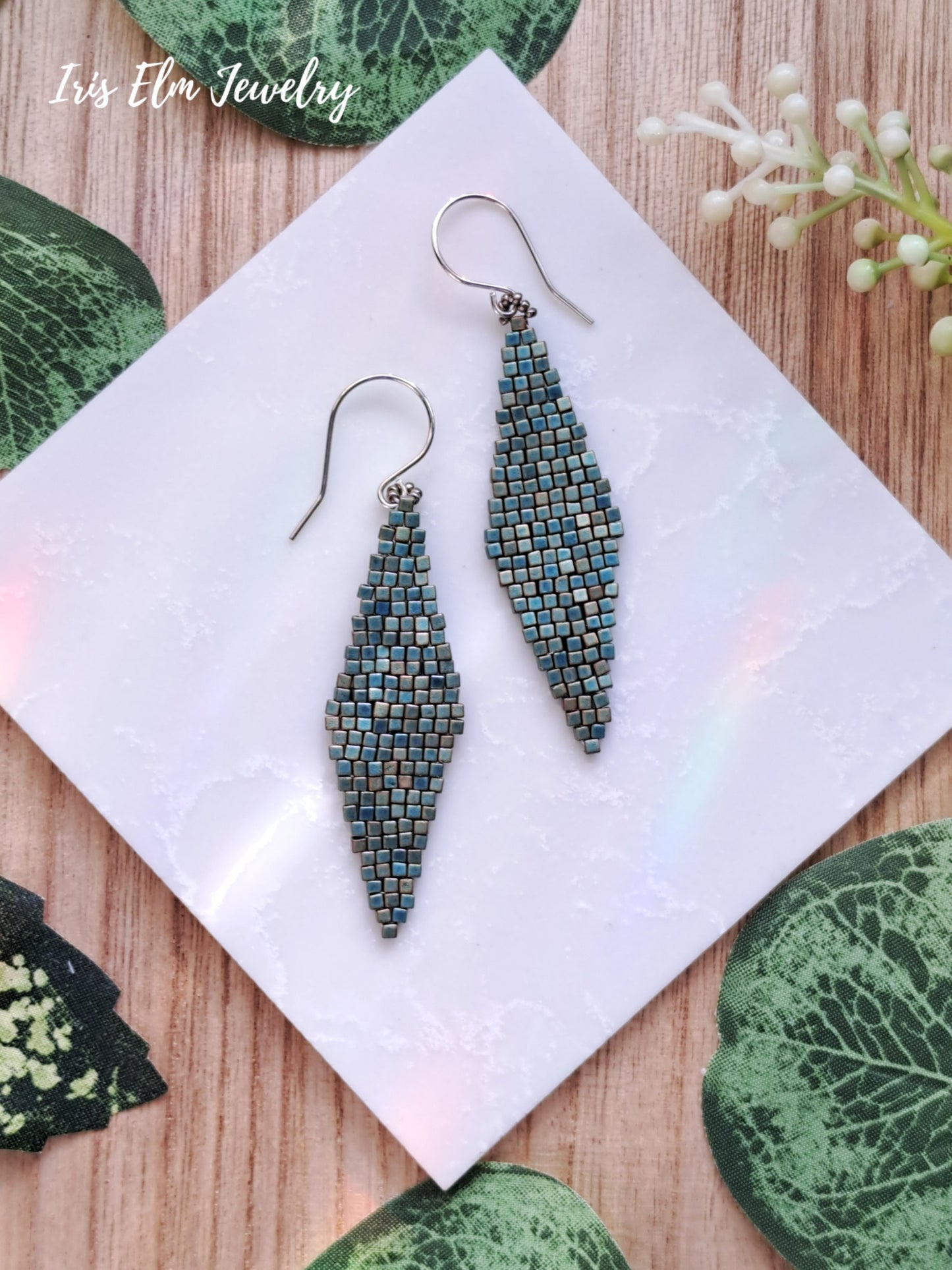 Pair of turquoise green matte iridescent diamond shape glass beaded earrings on a white napkin with leaf decorations.